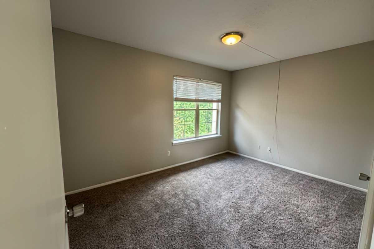A vacant, unfurnished room with light gray walls and a large window allowing natural light. The floor is covered with beige carpet, and there is a ceiling light fixture. A door is visible on the left side of the image, leading into the room. The overall atmosphere is neutral and empty, suggesting a space ready for new furnishings.