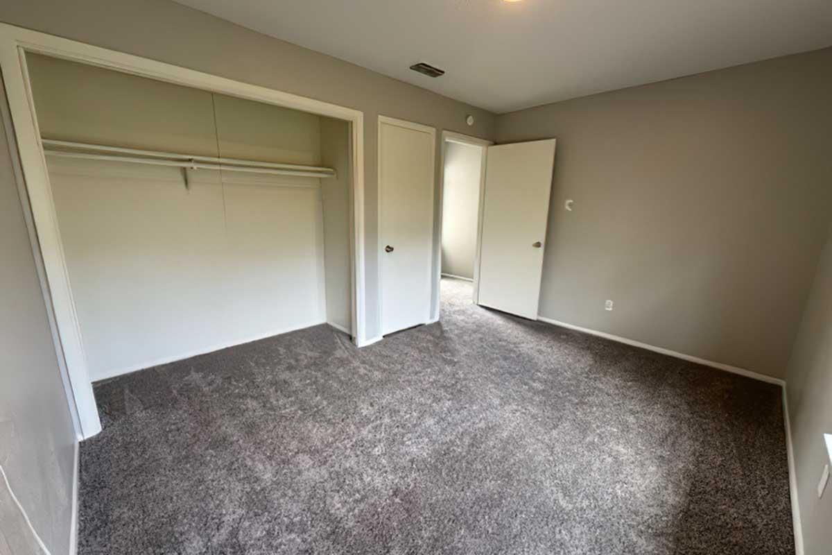 A vacant room featuring light gray walls, plush carpeted flooring, and a closet with an open door. Two closed doors lead to different areas, with one door partially open, revealing a glimpse of another space. The room is well-lit, showcasing a simple, clean aesthetic.