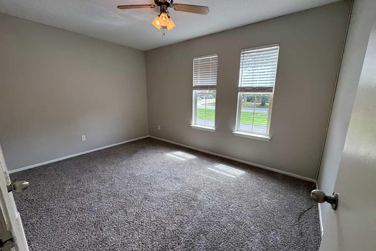 Empty room with a light gray carpet, featuring two windows letting in natural light. A ceiling fan is installed, and the walls are painted light gray. The door on the right is partially open, revealing a glimpse of a hallway outside.