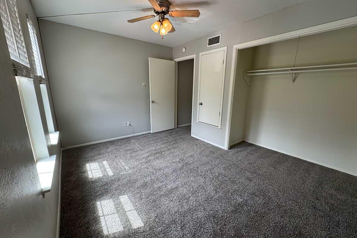A small, empty room with gray walls and carpet. It features a ceiling fan with light, a window allowing natural light, and a closet with a single shelf. The room appears clean and well-lit, perfect for a bedroom or office space.
