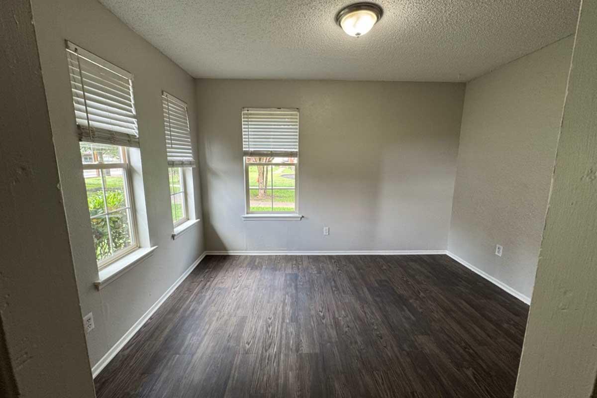Empty room with gray walls and a hardwood floor, featuring two windows with white blinds letting in natural light. A ceiling light fixture provides additional illumination. The space appears clean and ready for furnishing.