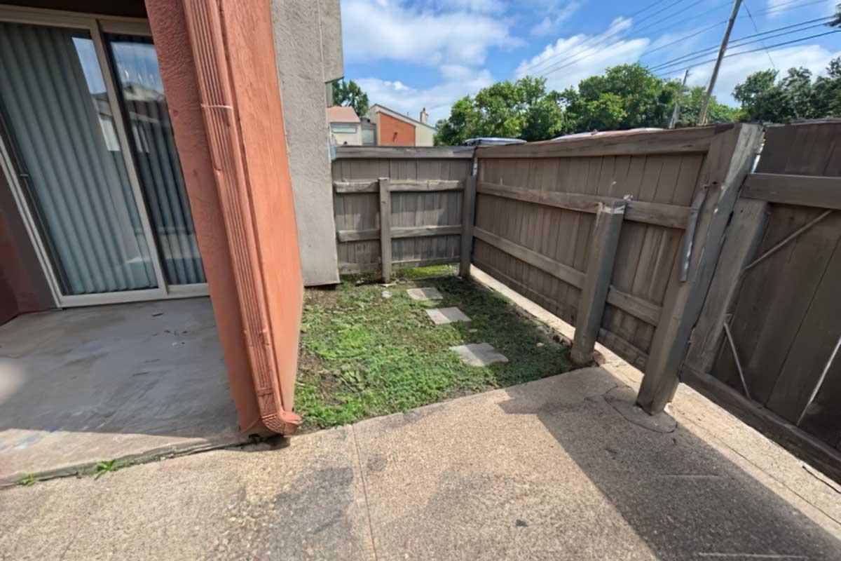 A small outdoor area next to a building, featuring a patch of grass and stepping stones. There is a wooden fence partially enclosing the space, and a wall of the building can be seen on the left. The sky is partly cloudy, and a few trees are visible in the background.