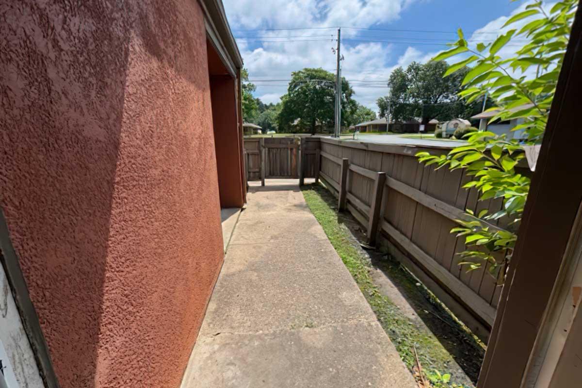 Narrow concrete pathway between two buildings, flanked by wooden fences. Grass grows along the edge. Clear blue sky with some clouds above. Sunlight casts shadows on the ground.