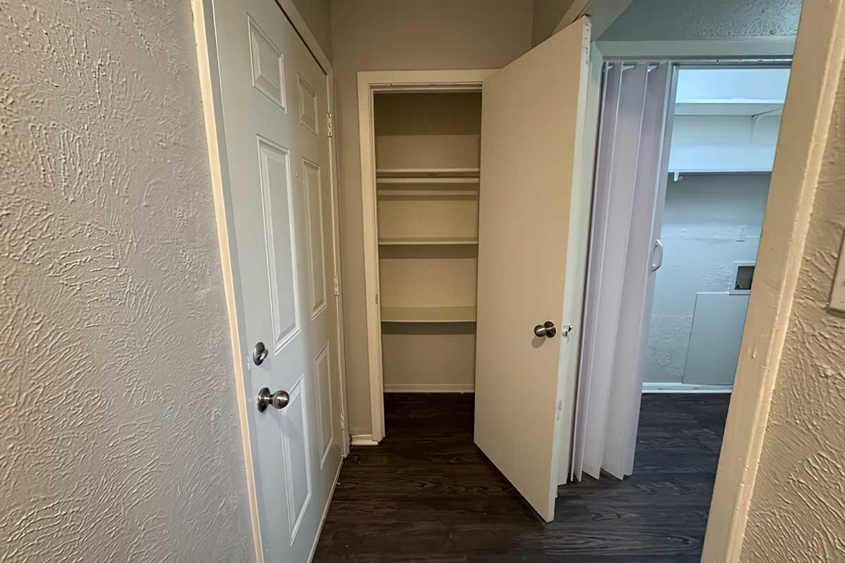 A small room with an open closet and a door leading to another space. The walls are light-colored, and the flooring is dark wood. Shelves are visible inside the closet. There are two doors, one of which is partially open, providing a glimpse into a different area.