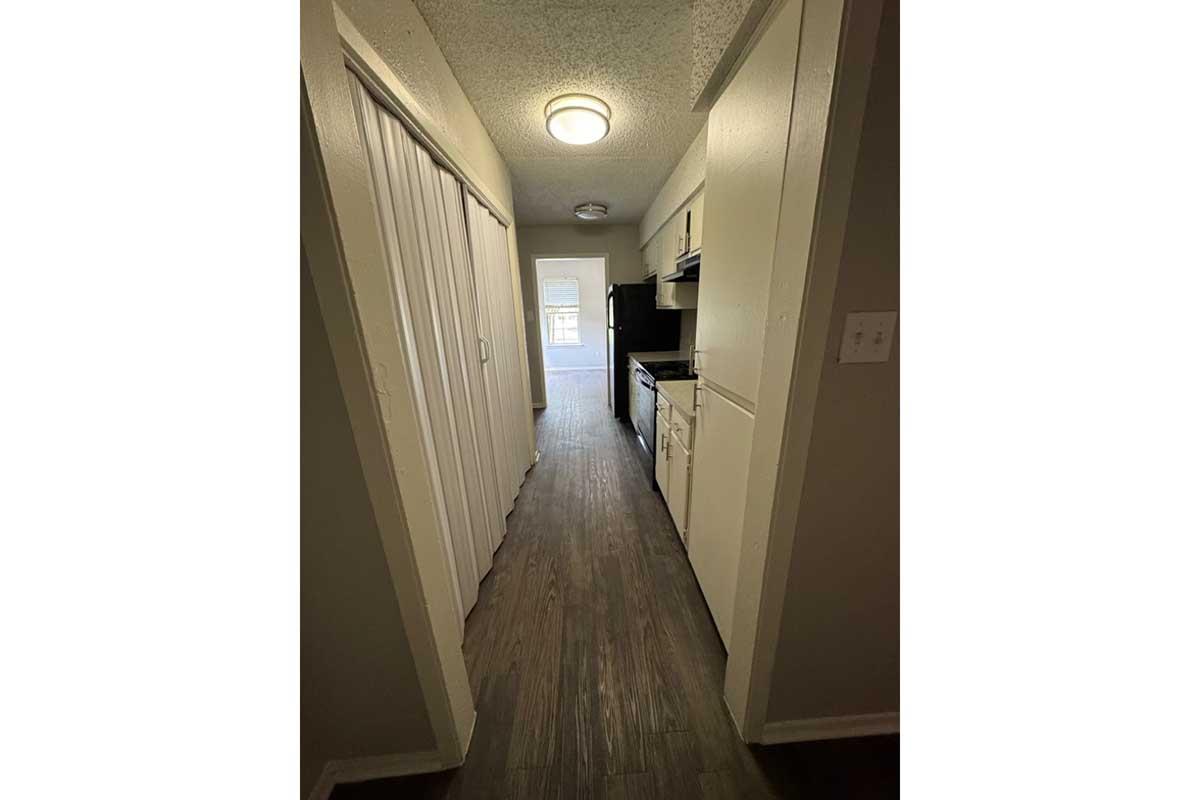 A narrow hallway leading to a kitchen area. The kitchen features white cabinets, a black refrigerator and stove, and a window at the far end, allowing natural light. The flooring is dark wood, and the walls are painted in neutral colors, creating a clean and modern appearance.