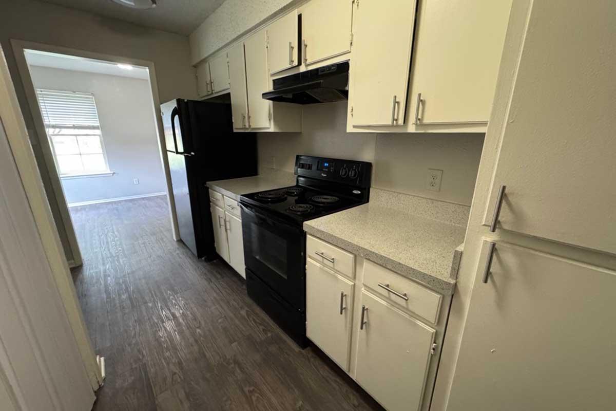 A modern kitchen featuring white cabinetry, a black refrigerator, and a black stove with an oven. The countertops are light-colored, and the flooring is wood-like. There is a window providing natural light from a nearby room. The overall design is clean and functional.