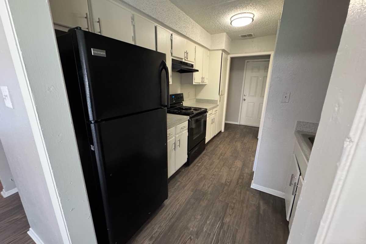 A modern kitchen featuring black appliances, including a refrigerator and stove, with white cabinets and gray countertops. The room has a light-colored wall and wood-like flooring, with a doorway leading to another area. The space is well-lit by overhead lights.