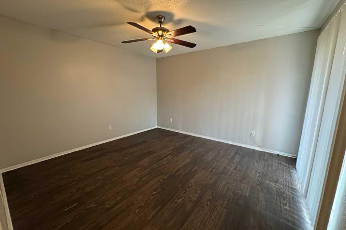 An empty room with light brown wooden floors, a ceiling fan with four light bulbs, and light gray walls. There is a window on one side with sheer curtains, allowing natural light to fill the space. The room appears spacious and unfurnished, ready for decoration or use.