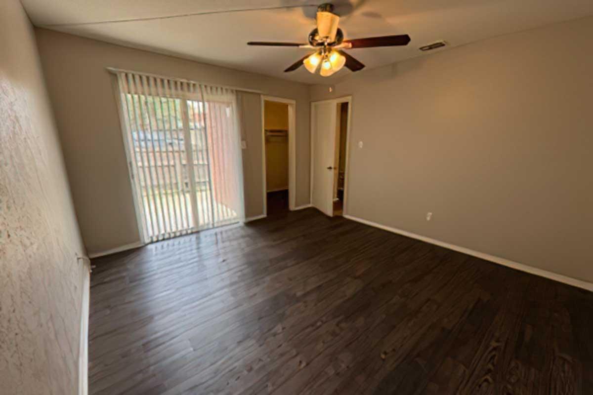A spacious, empty room with dark wood flooring, a ceiling fan, and a sliding glass door leading to a patio. The walls are painted a light neutral color, and there is a closet with an open door in the background. Natural light is coming in from outside.