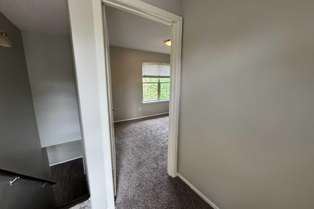 A hallway leading to a room with light-colored walls and a large window. The floor is carpeted, and a staircase is visible on the left. Natural light brightens the space, creating a welcoming atmosphere.