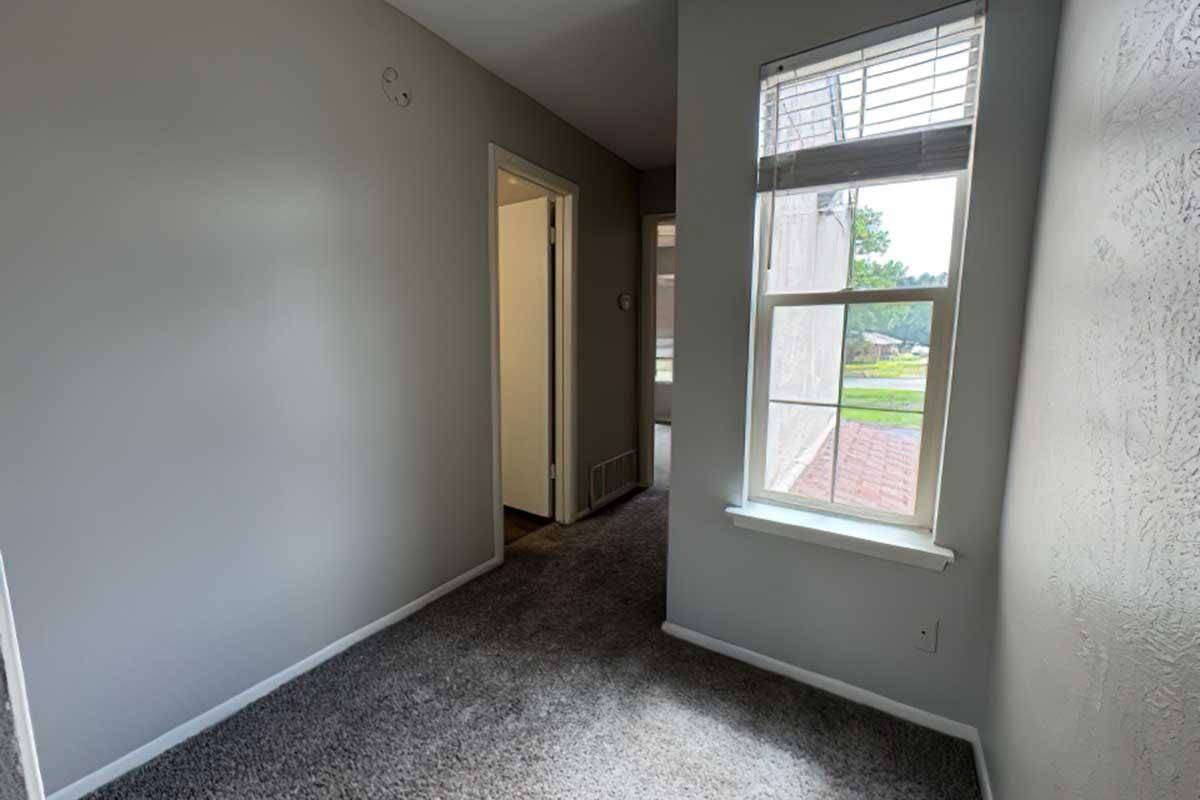 Interior view of a hallway with light gray walls and carpet. A window provides natural light, and a door leads to another room. The space appears well-lit and clean, featuring a simple and modern design.