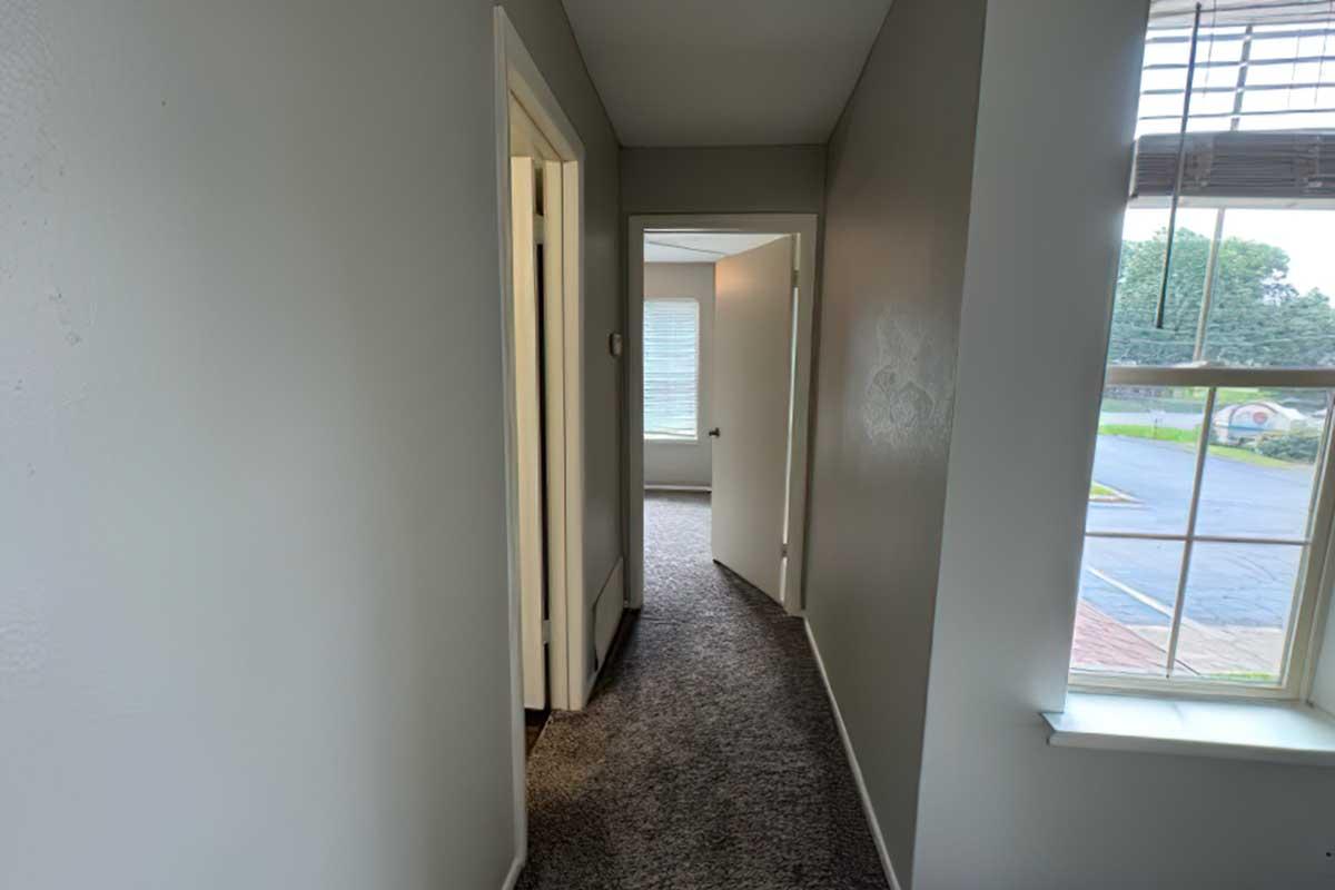 A narrow hallway in a light-colored room, with several doorways leading to unknown rooms. The flooring is carpeted, and a window at the end offers natural light. The walls are painted in a neutral tone.