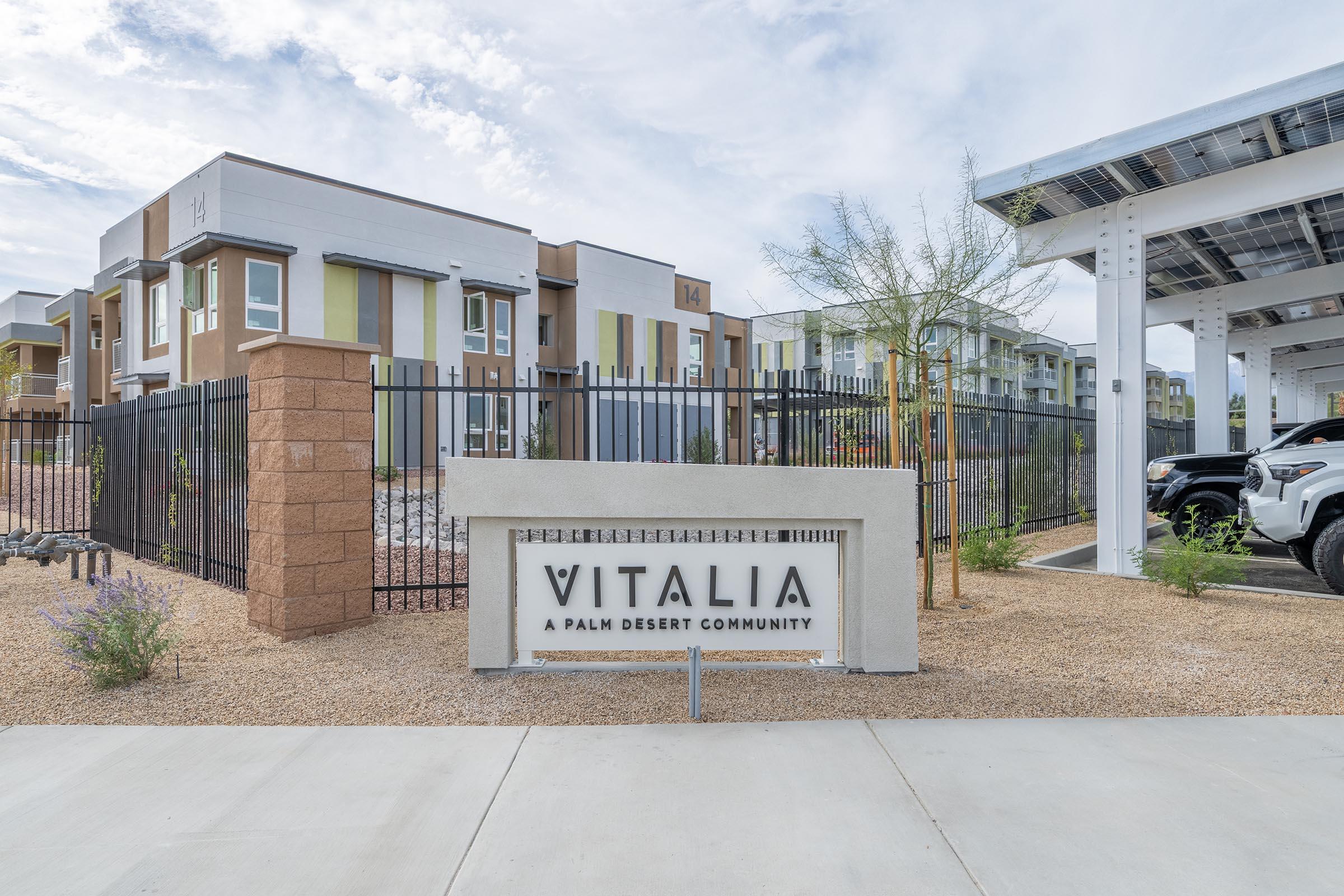 Sign for "Vitalia, a Palm Desert Community," prominently displayed in front of a modern residential building. The building features contemporary architecture with multiple stories, surrounded by desert landscaping and a fence. In the background, there are solar panels and parked vehicles.