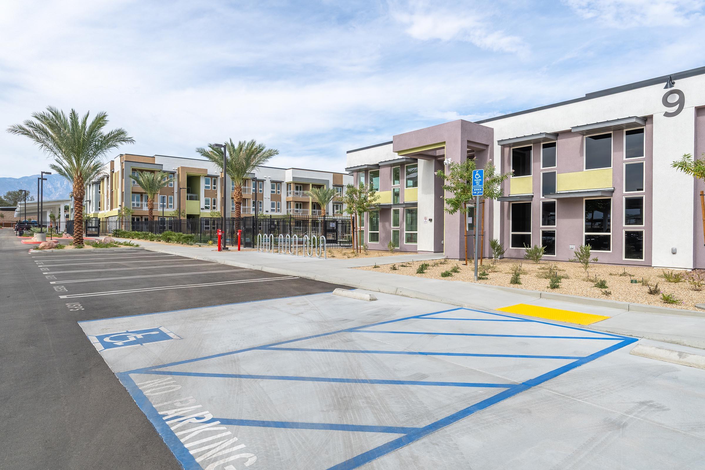 Modern apartment complex with multiple buildings. The foreground features designated parking spaces, including one labeled for handicapped access, and two fire hydrants. Palm trees line the sides of the paved area, with bike racks visible in the background and landscaped gardens adding greenery to the setting.