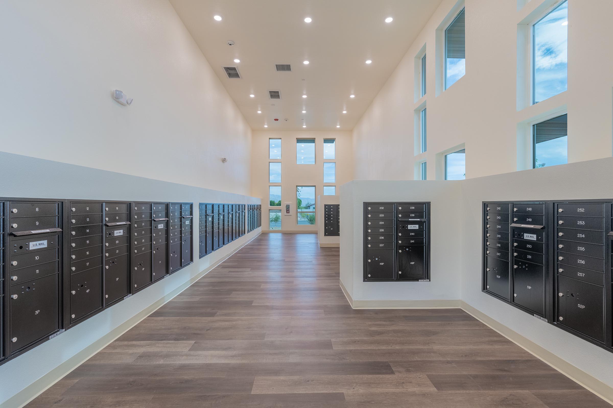 A modern indoor mailroom featuring multiple rows of secure mailboxes along the walls. The space is well-lit with large windows, high ceilings, and a polished wooden floor, creating a clean and organized environment for residents to access their mail.
