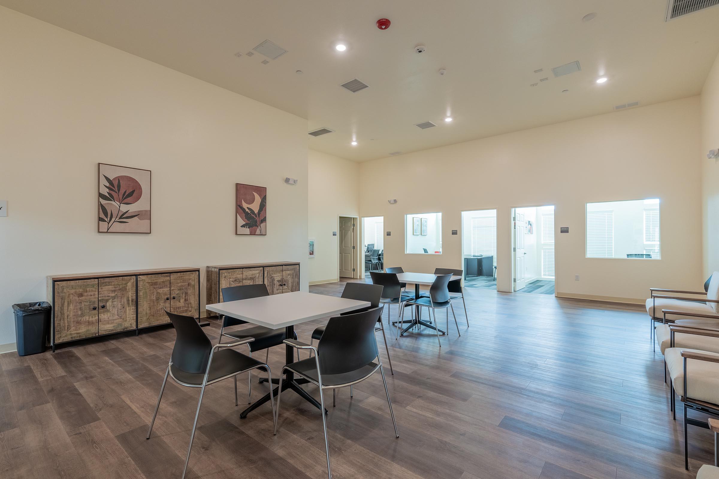 A spacious room featuring a few tables and chairs, artwork on the walls, and wooden cabinets. The flooring is wood-like, and there is a doorway leading to another area. The space is well-lit with a modern, minimalist design.