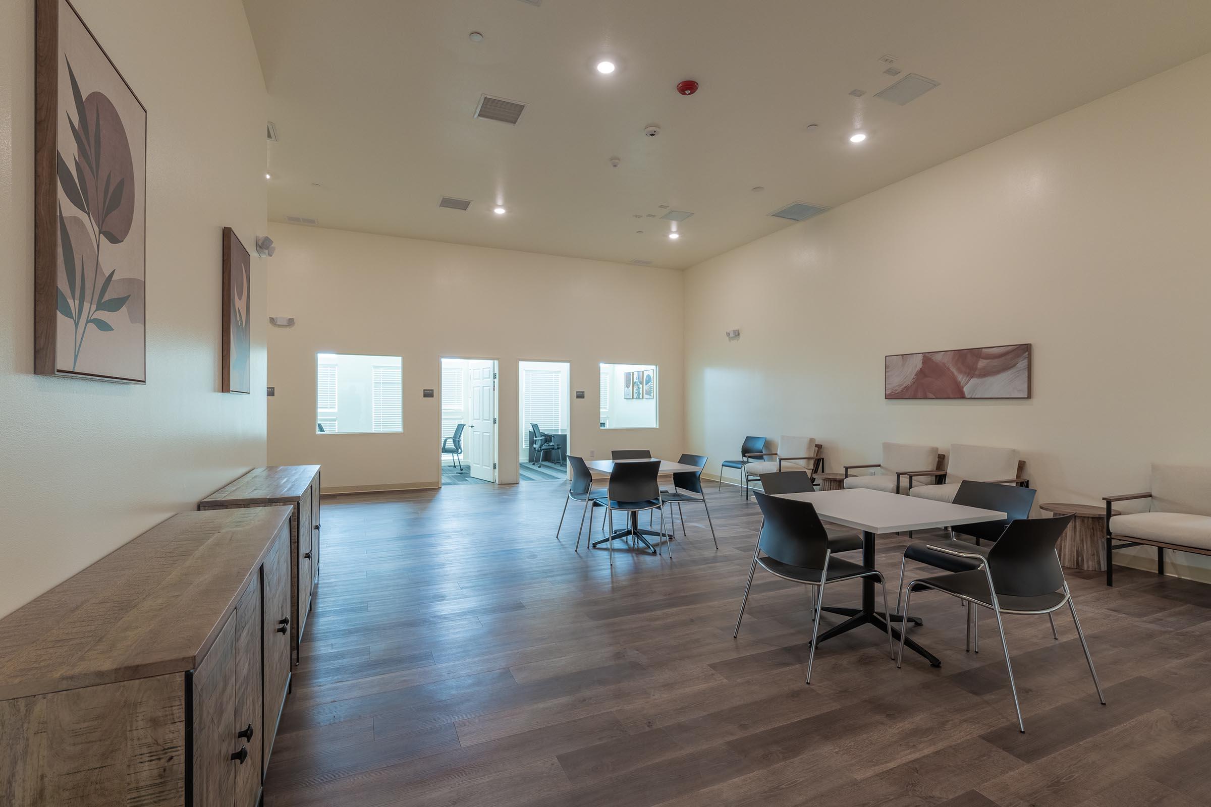 A spacious, modern interior room with light-colored walls and wooden flooring. It features a circular dining table with black chairs and several beige armchairs arranged around the space. There are two doors leading to other areas, and decorative wall art enhances the aesthetic.