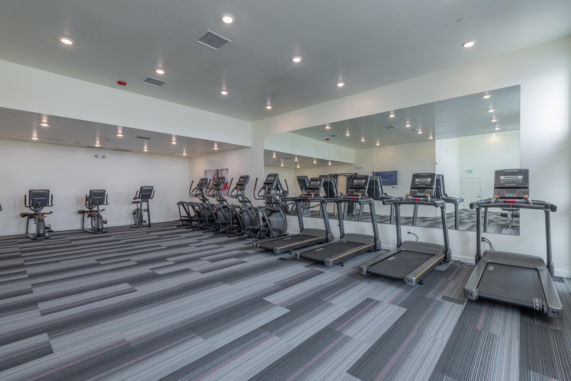 A modern fitness room featuring several treadmills and elliptical machines, with a large mirror on one wall and well-lit, neutral-colored walls. The floor has a gray and red striped carpet, creating a contemporary workout environment.