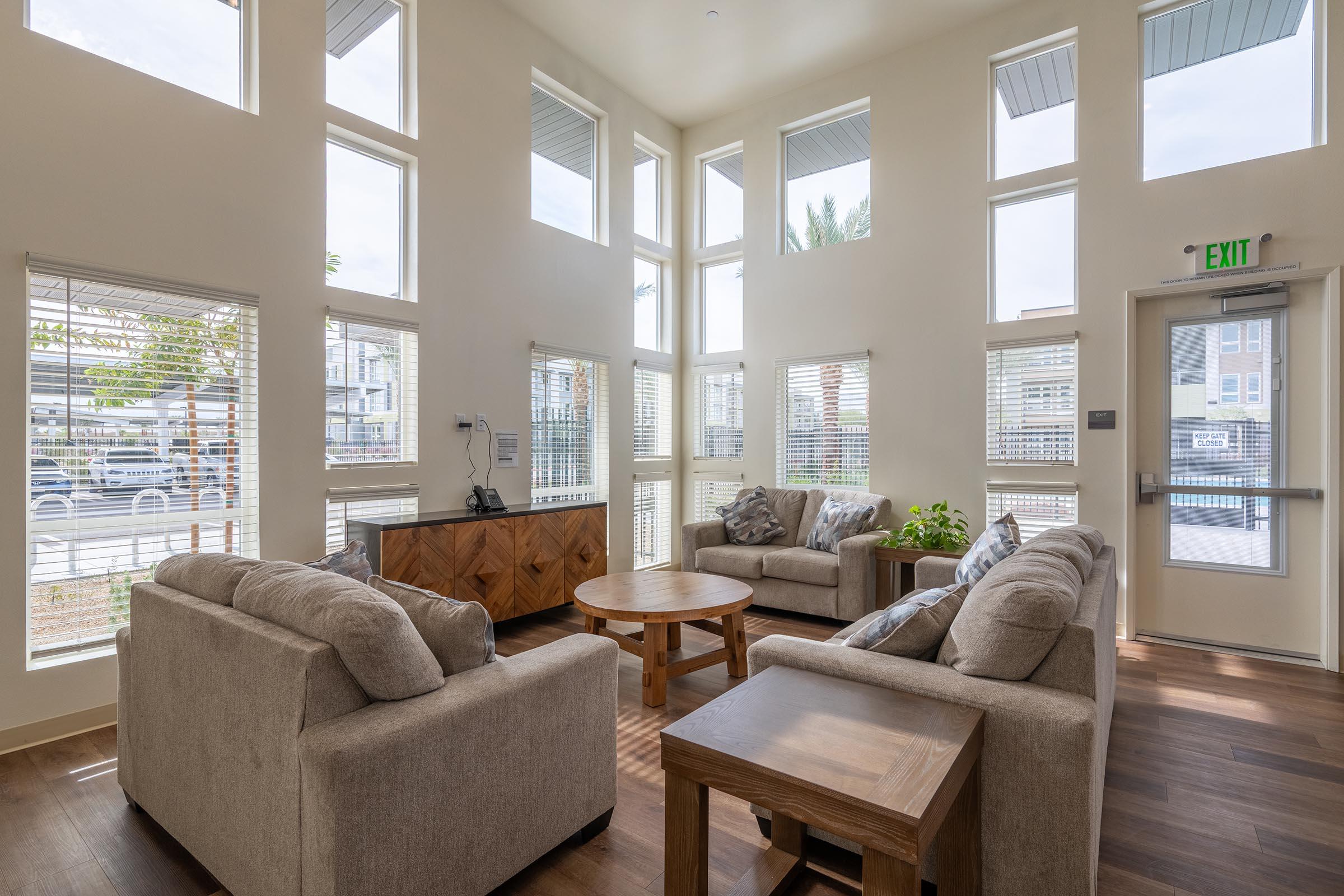 A modern interior of a bright living space featuring large windows, a cozy seating arrangement with two beige sofas, a round wooden coffee table, and decorative plants. Natural light fills the room, creating an inviting atmosphere. An exit door and wooden cabinet are visible in the background.