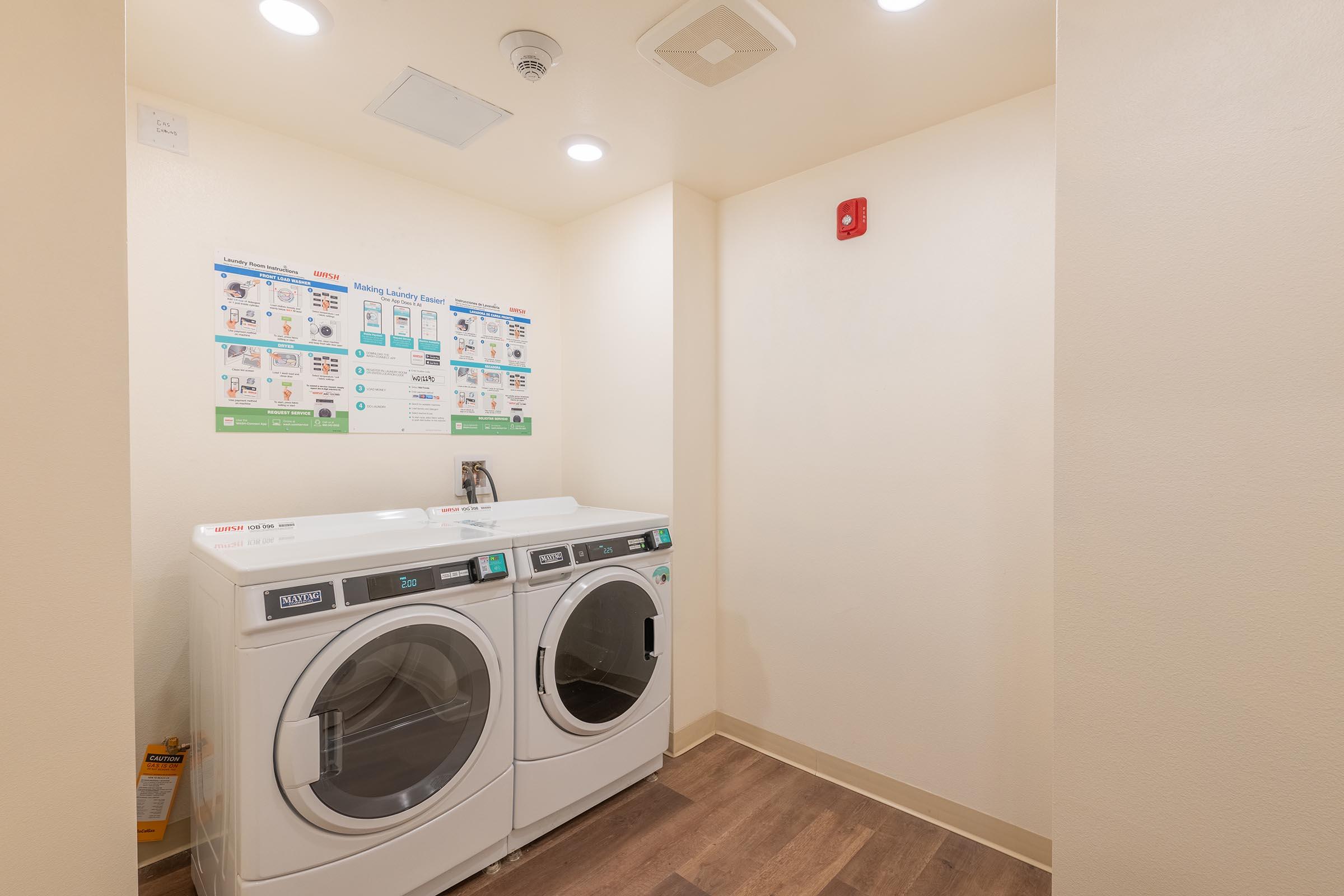 A clean laundry room featuring a stackable washer and dryer in a bright, well-lit space. The walls are painted in a light color, and there's a bulletin board with laundry instructions and tips. The floor is made of wood laminate, enhancing the modern look of the room.