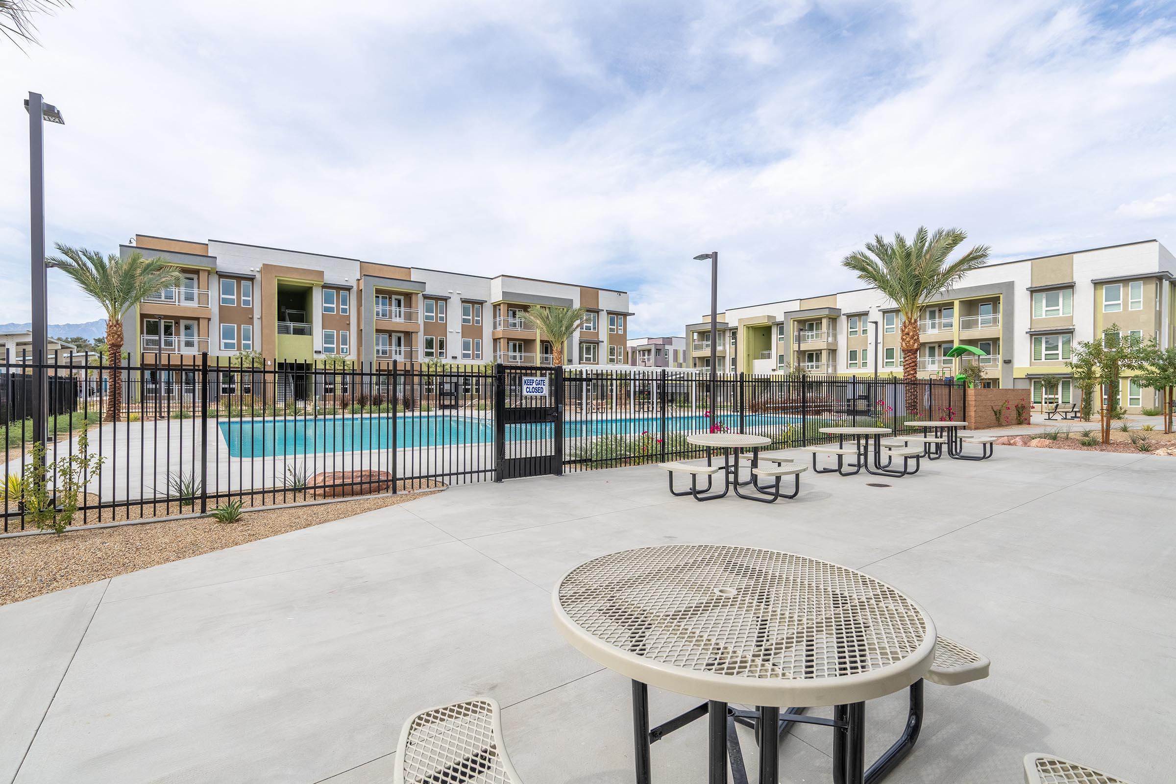 A modern apartment complex featuring a swimming pool enclosed by a black fence. There are several palm trees around the pool area, and picnic tables are set up on a concrete patio in the foreground. The sky is partly cloudy, and the buildings are painted in neutral tones.