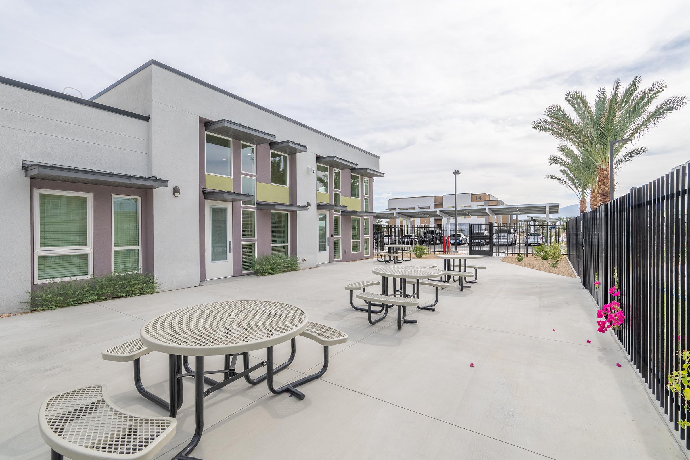 Outdoor area featuring several metal picnic tables on a concrete surface, surrounded by green plants. A modern building with large windows and a palm tree is visible in the background, along with a covered parking area. The overall setting appears inviting and well-maintained.