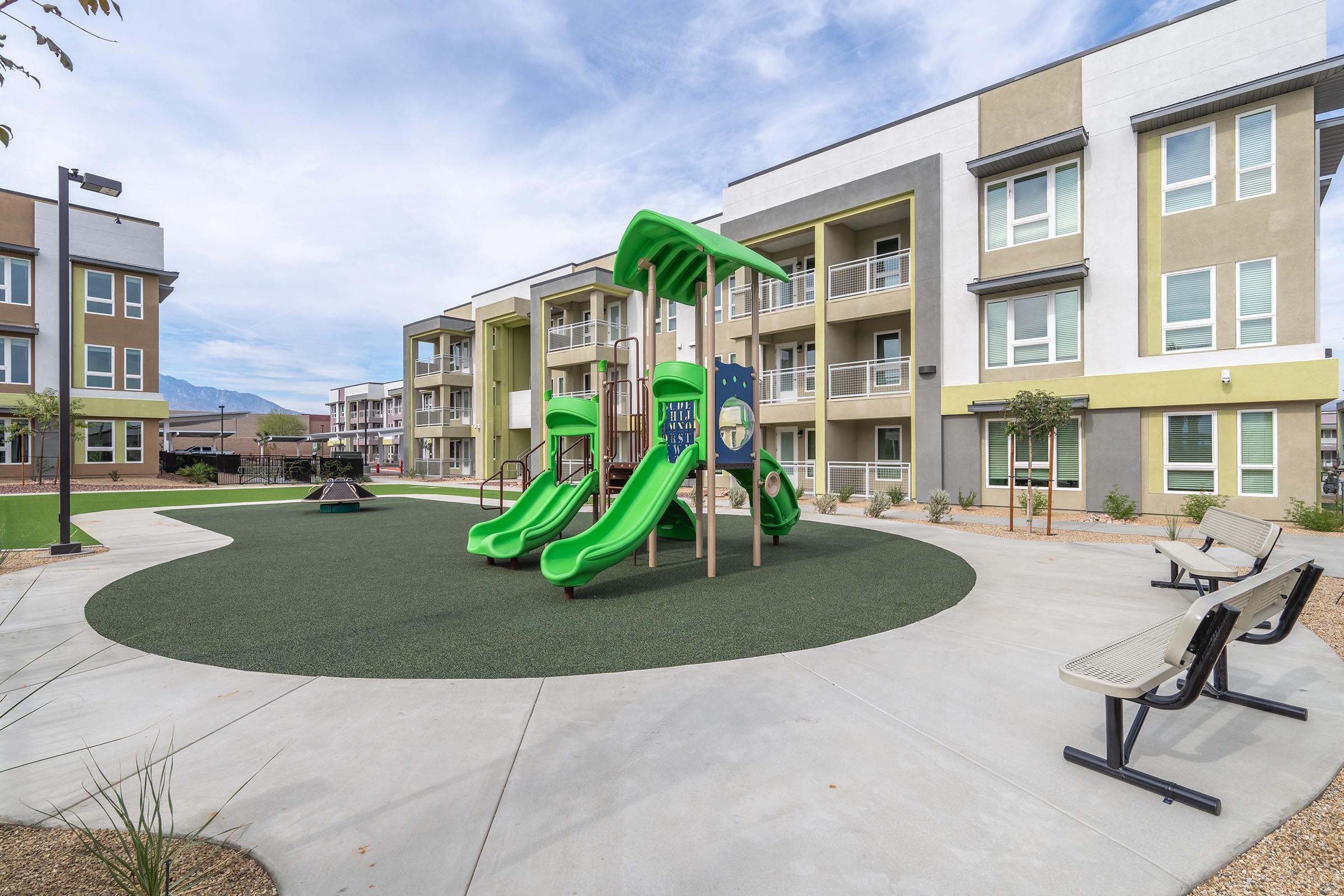 A modern playground with green slides and climbing equipment, surrounded by a landscaped area featuring benches. In the background, there are multi-story residential buildings under a clear blue sky. The playground is designed for children and is situated in a well-maintained outdoor space.