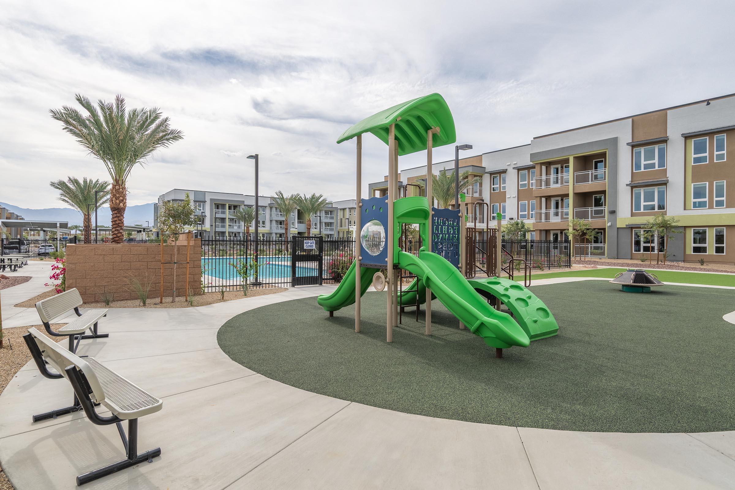 Playground area featuring a green play structure with slides, surrounded by turf. Benches are placed nearby for seating. In the background, there are modern apartment buildings and a swimming pool, with palm trees adding a tropical touch to the landscape.