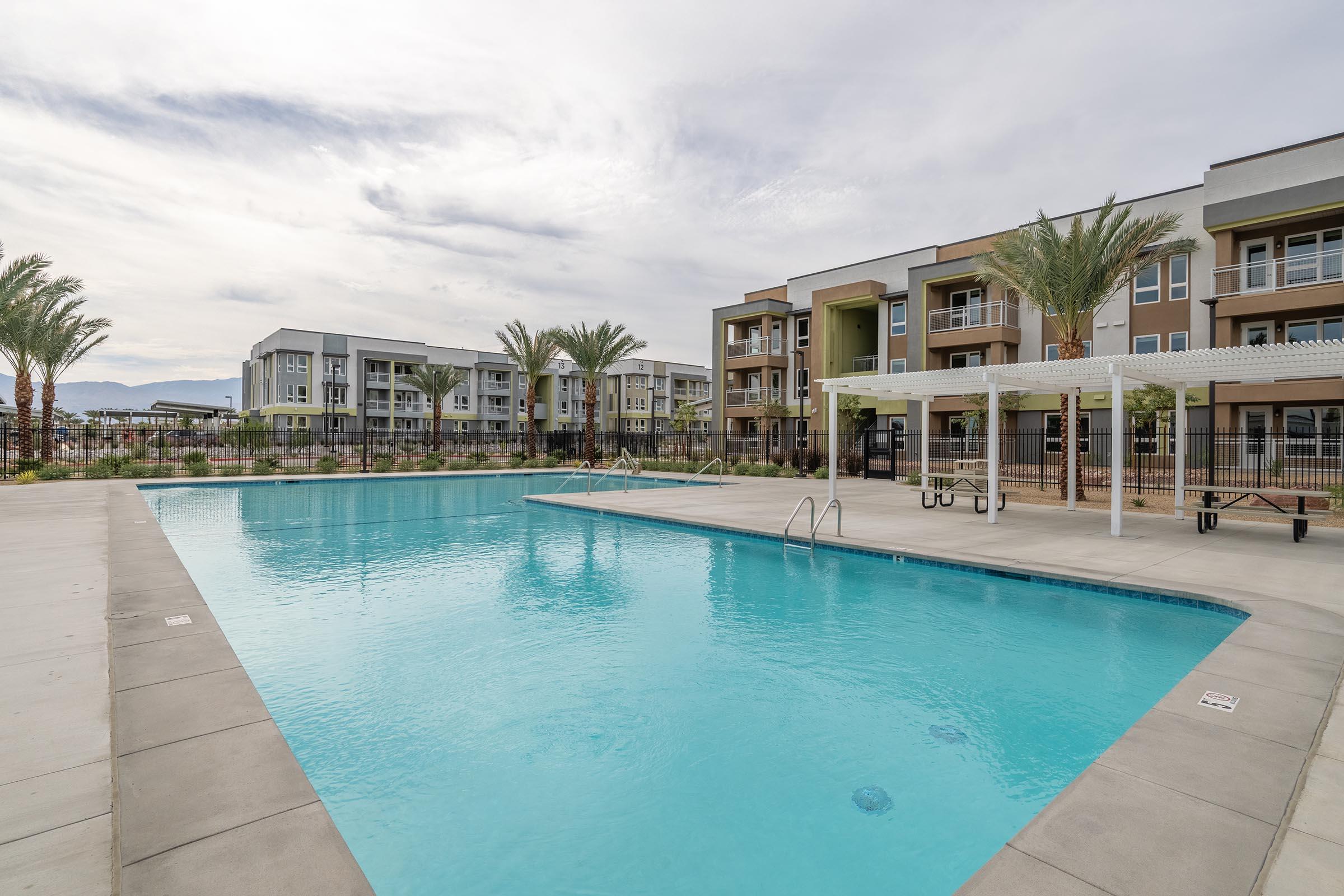 A clear blue swimming pool surrounded by palm trees and modern apartment buildings. The pool area features lounge chairs and shaded seating, set against a cloudy sky. The complex showcases contemporary architecture with balconies and landscaped greenery.