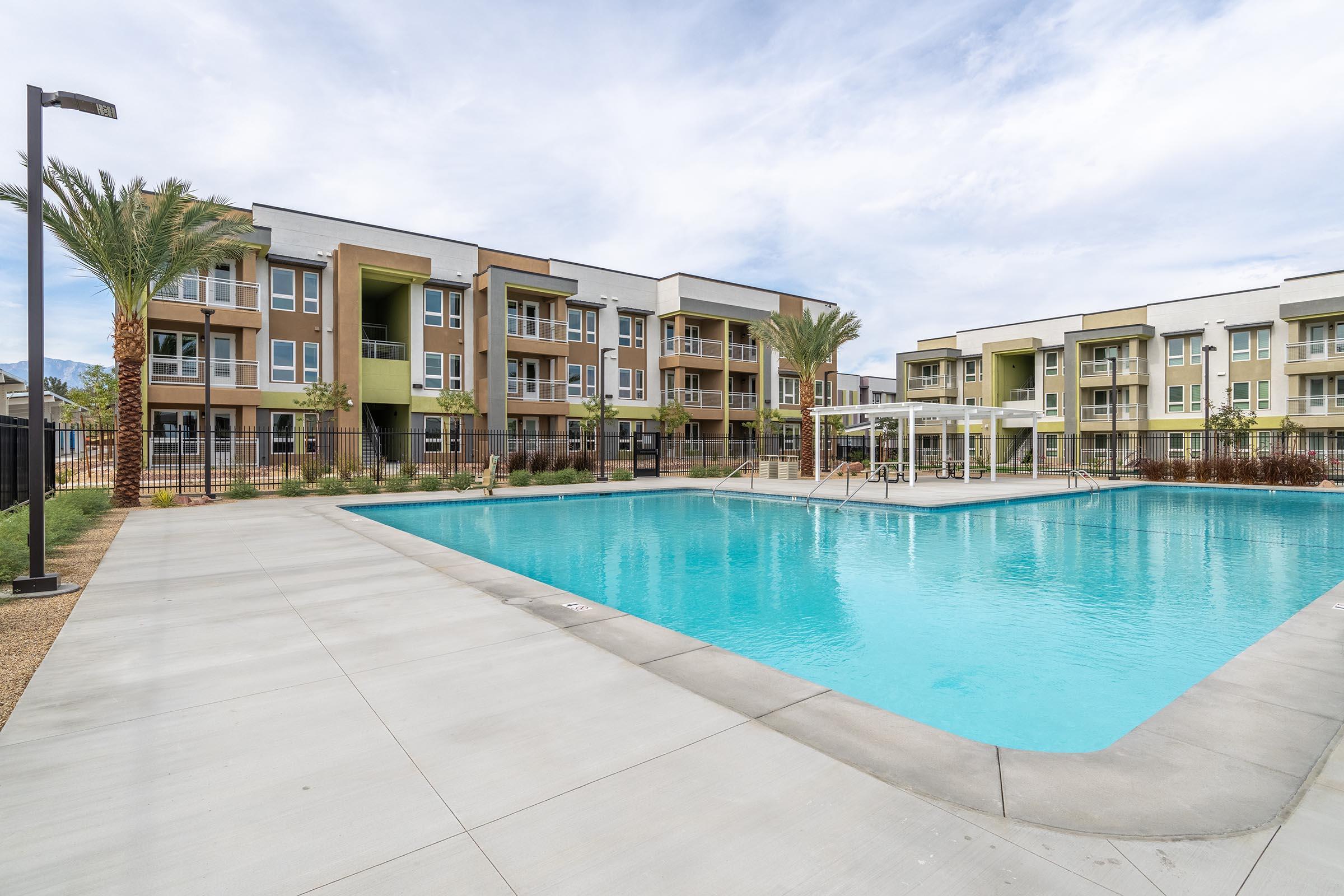 A modern apartment complex featuring two buildings with balconies overlooking a clear blue swimming pool. Palms and landscaped greenery surround the pool area, which includes lounge chairs and a shaded seating area. The sky is partly cloudy, creating a pleasant atmosphere for leisure.