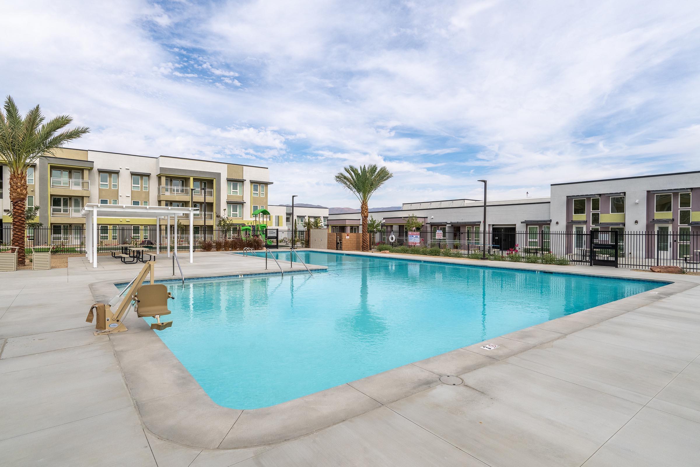 A sunny outdoor swimming pool surrounded by palm trees and modern apartment buildings. The pool features a diving board and lounge chairs. Clear blue skies reflect above, creating a tranquil atmosphere for relaxation and leisure. The area is well-maintained with a shaded seating area nearby.