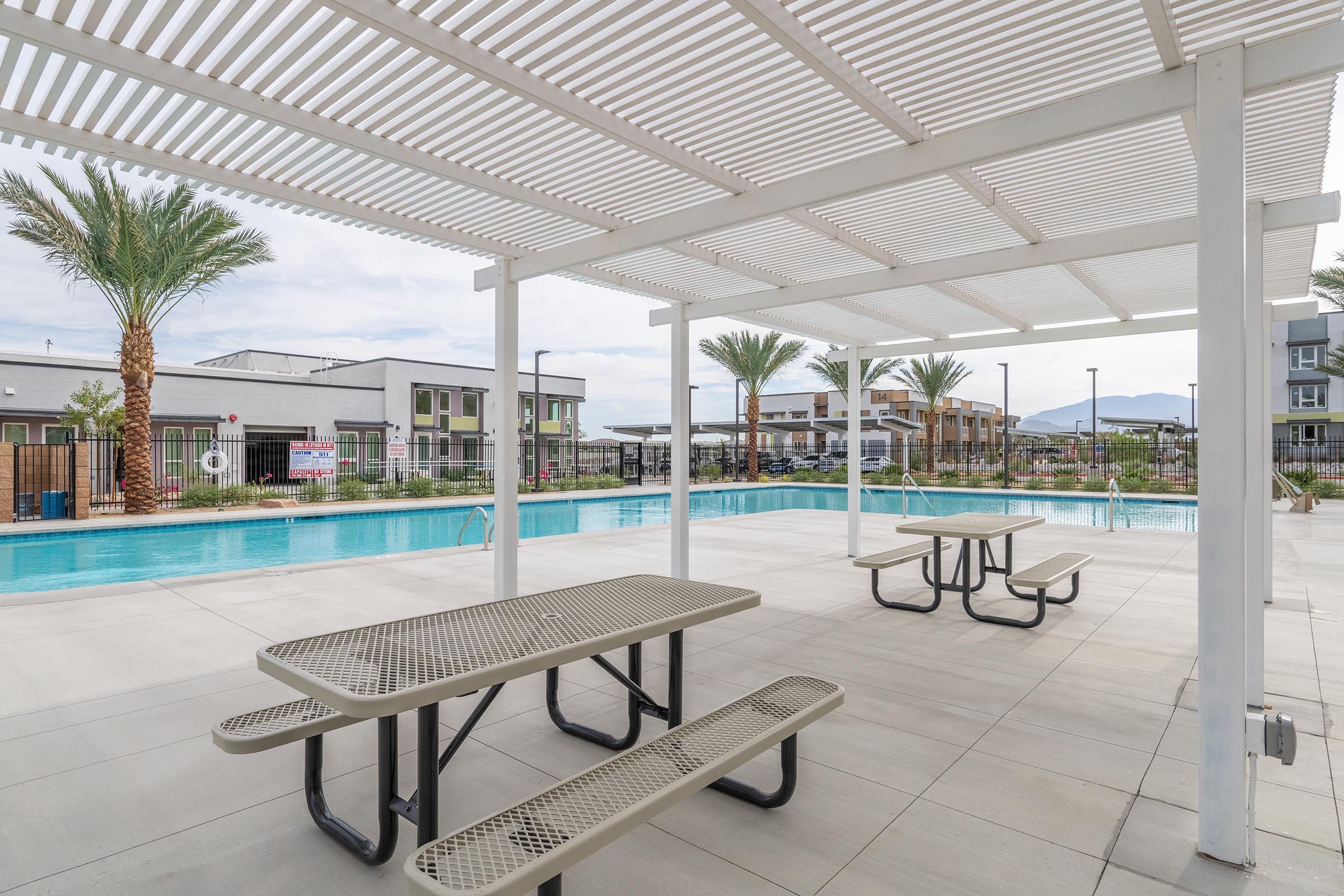 A spacious outdoor area featuring a covered seating section with picnic tables beside a swimming pool. Palm trees are visible in the background, along with residential buildings. The sky is partly cloudy, creating a relaxing atmosphere for outdoor activities.