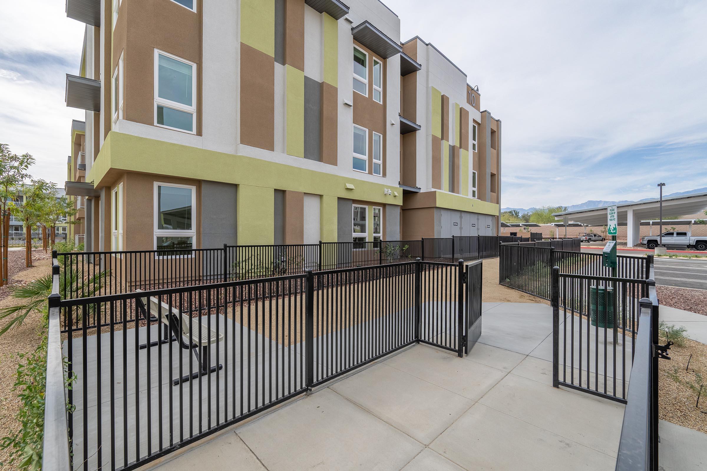 A modern multi-story building with a mix of brown, green, and beige exterior colors. A fenced entrance leads to a landscaped area, with concrete pathways and seating. The background shows a parking lot and additional buildings, under a cloudy sky.