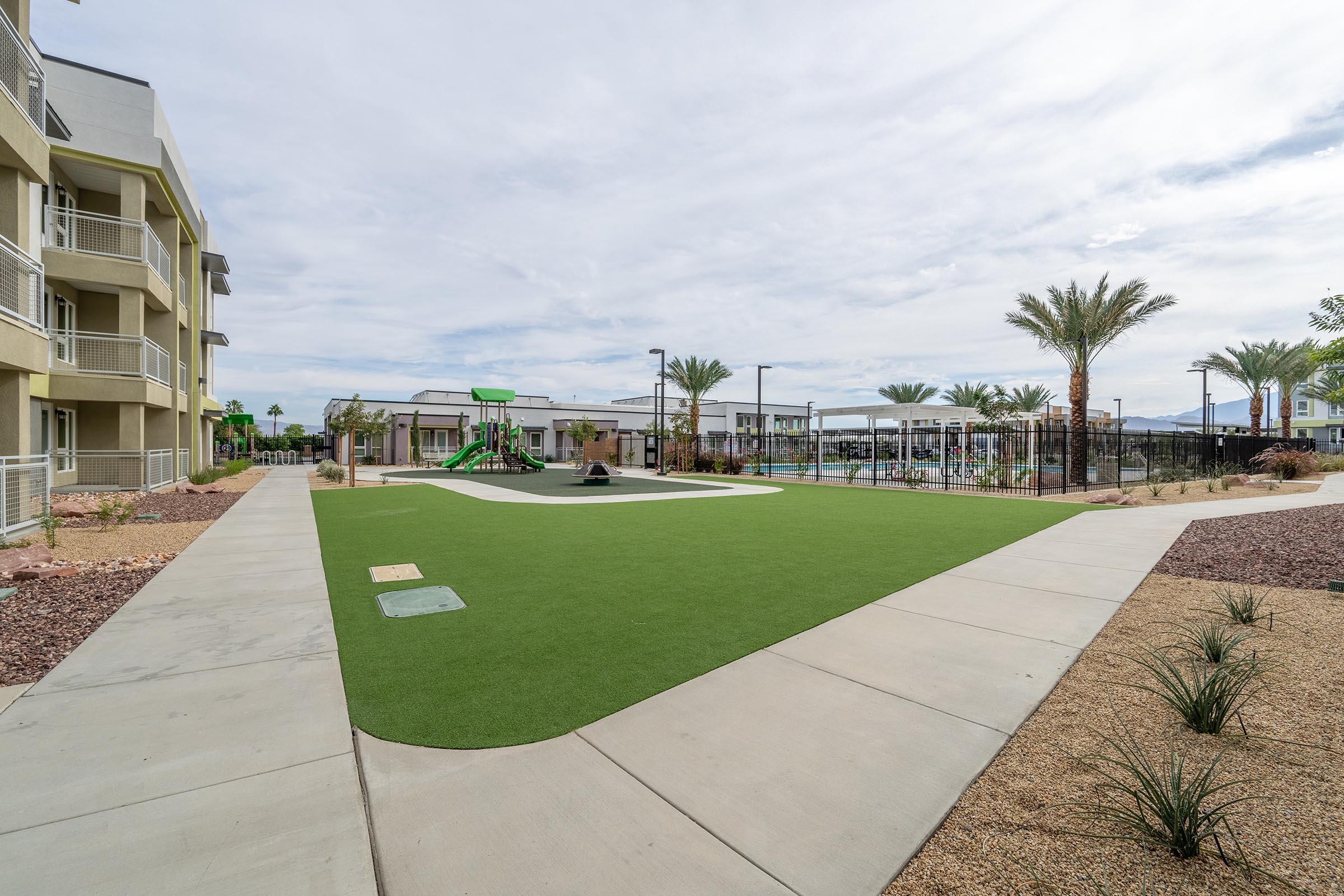 A spacious outdoor area featuring artificial grass, pathways, and palm trees. In the background, there are playground equipment and a swimming pool area. Nearby buildings provide a modern residential environment. The sky is partly cloudy, creating a pleasant atmosphere for outdoor activities.