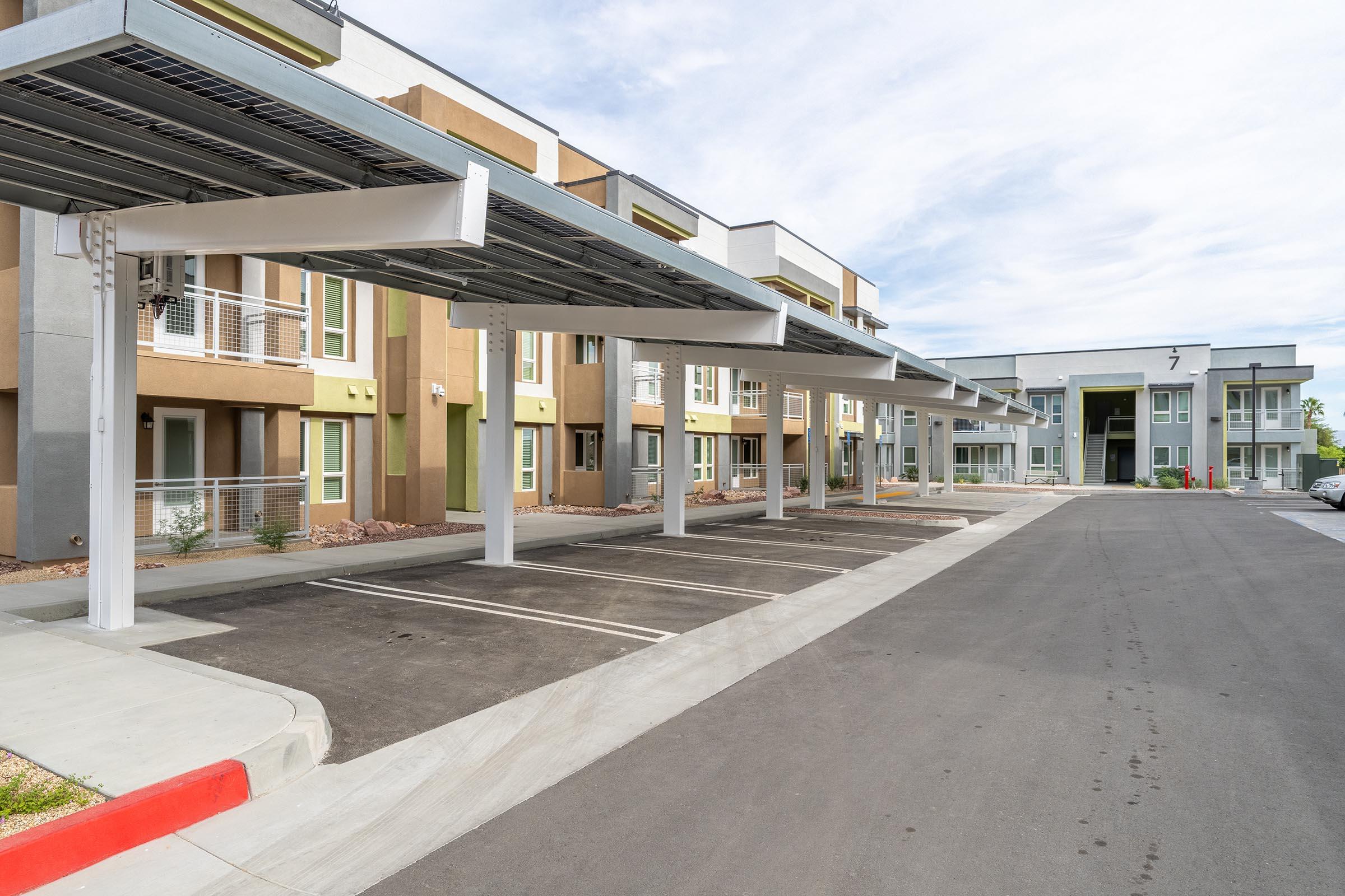 A modern apartment complex featuring several buildings with balconies and colorful facades. Covered parking spaces line the front, with a smooth asphalt road leading to the entry. The sky is partly cloudy, giving a bright and inviting atmosphere to the area.