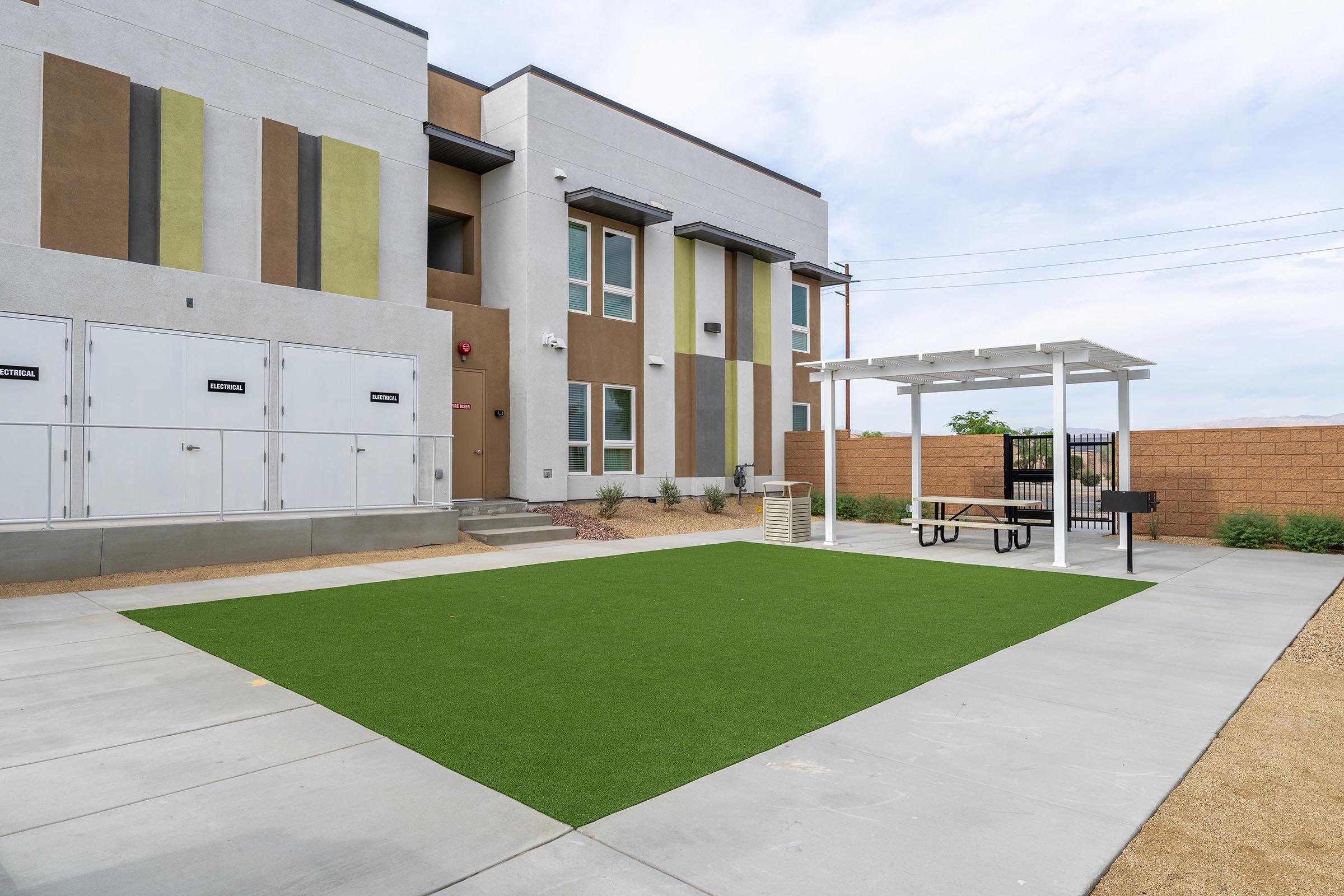 A view of a modern building exterior featuring a green artificial lawn area with a picnic table and a gazebo. There are storage units nearby, and the landscape is simple with gravel and low-maintenance plants. The sky is partly cloudy, contributing to a serene outdoor setting.