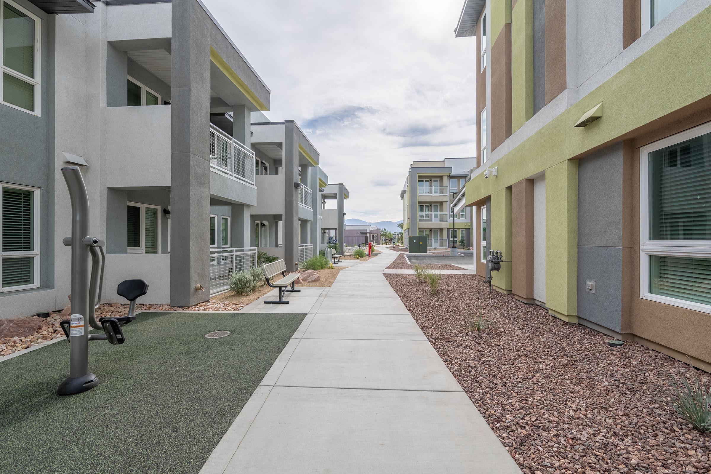 A modern residential complex featuring two-story buildings with varied color schemes. A paved pathway runs through the center, flanked by landscaped areas and exercise equipment. The sky is partly cloudy, and the surroundings are clean and well-maintained, creating an inviting atmosphere for residents.
