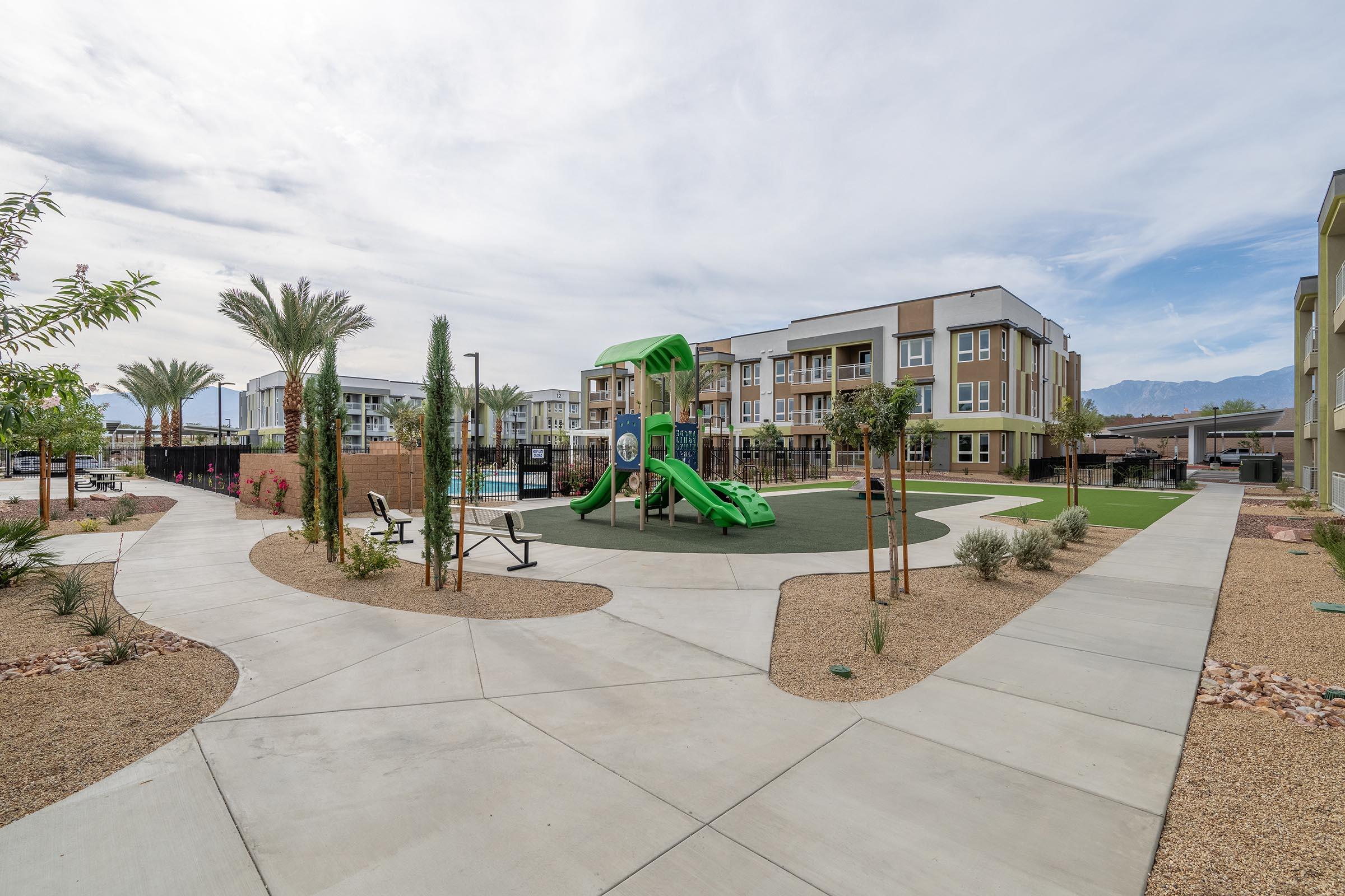 A modern residential area featuring a playground with a green slide and play structures, surrounded by landscaped pathways, palm trees, and picnic benches. The background shows contemporary apartment buildings against a partly cloudy sky.