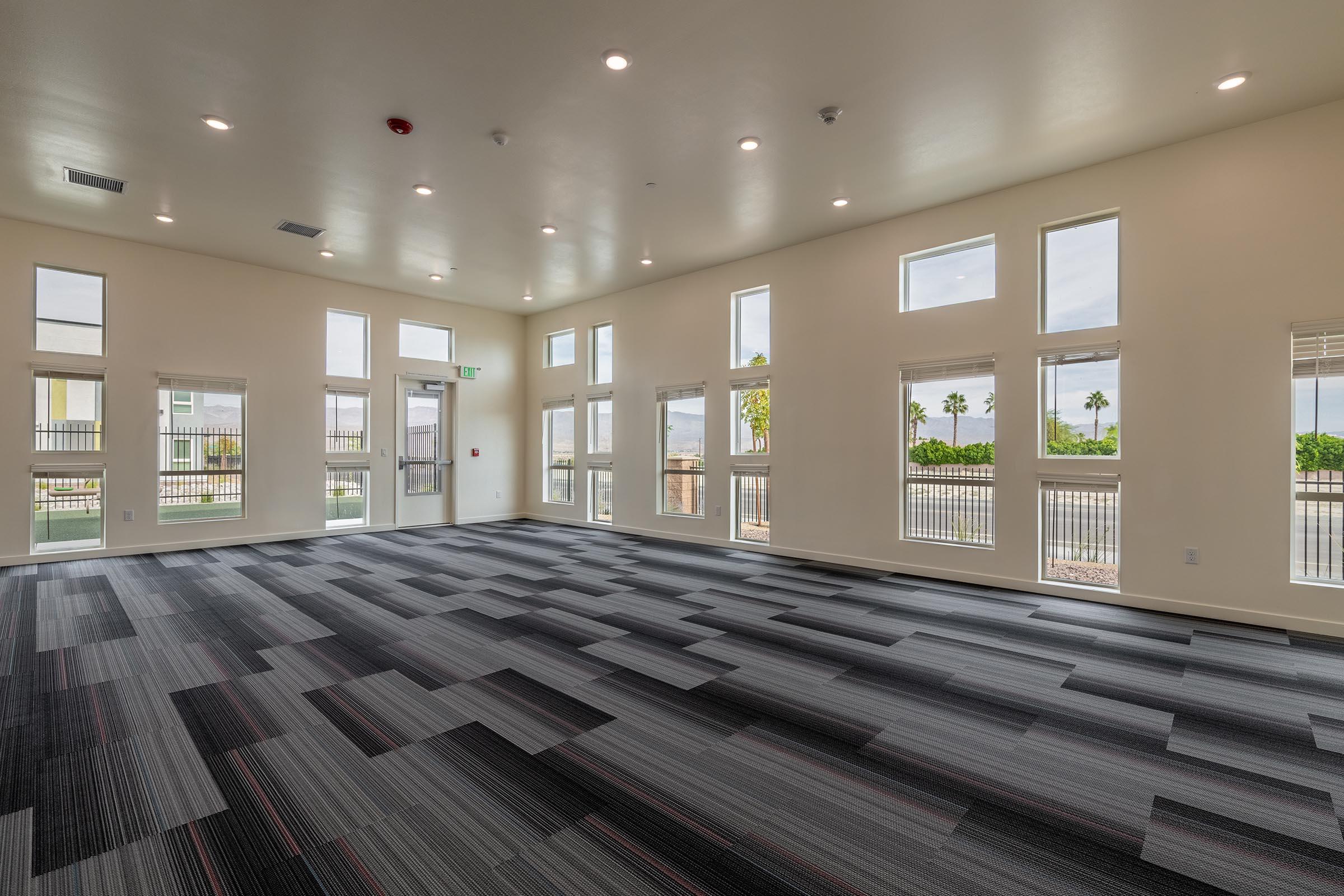 A spacious, well-lit room featuring multiple windows, allowing natural light to fill the space. The floor is covered with modern patterned carpeting in shades of gray and black. The walls are painted white, creating a clean, bright atmosphere. Ideal for meetings or gatherings.