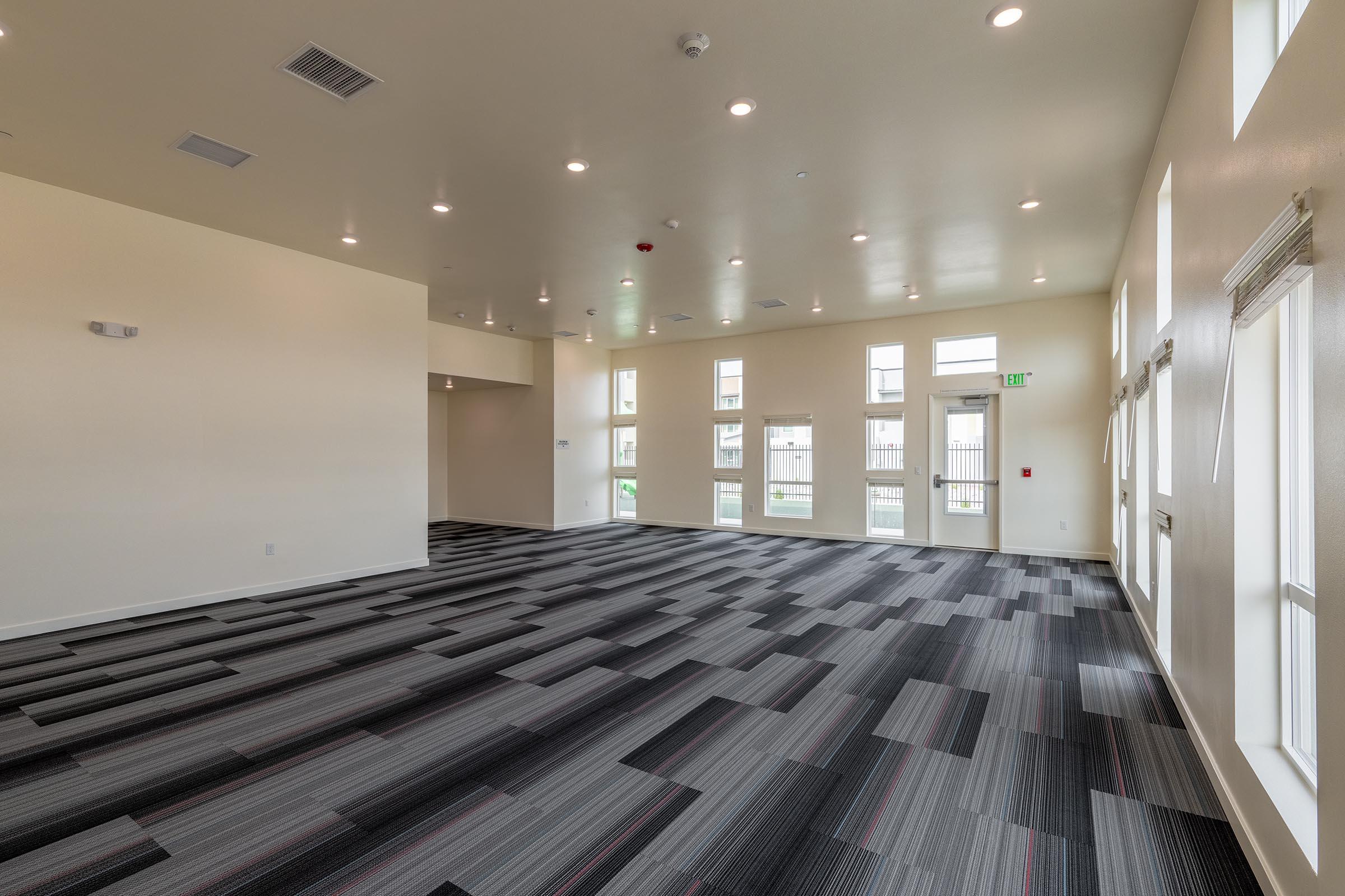 A spacious, well-lit interior room with large windows and a modern design. The floor features a striped carpet in gray and black tones. The walls are painted a light color, and the ceiling has recessed lighting. There are no furnishings, giving the space a clean, open feel.