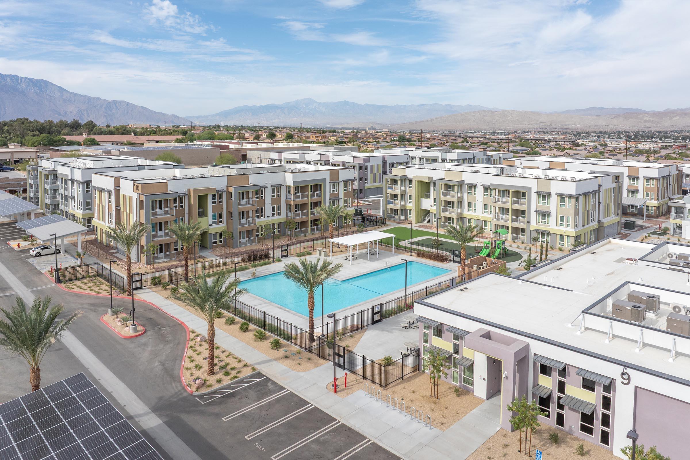 Aerial view of a modern residential complex featuring multiple buildings with light-colored exteriors. In the foreground, there's a swimming pool surrounded by palm trees, with children's play equipment nearby. The background shows mountainous terrain and a clear blue sky, indicating a sunny day.