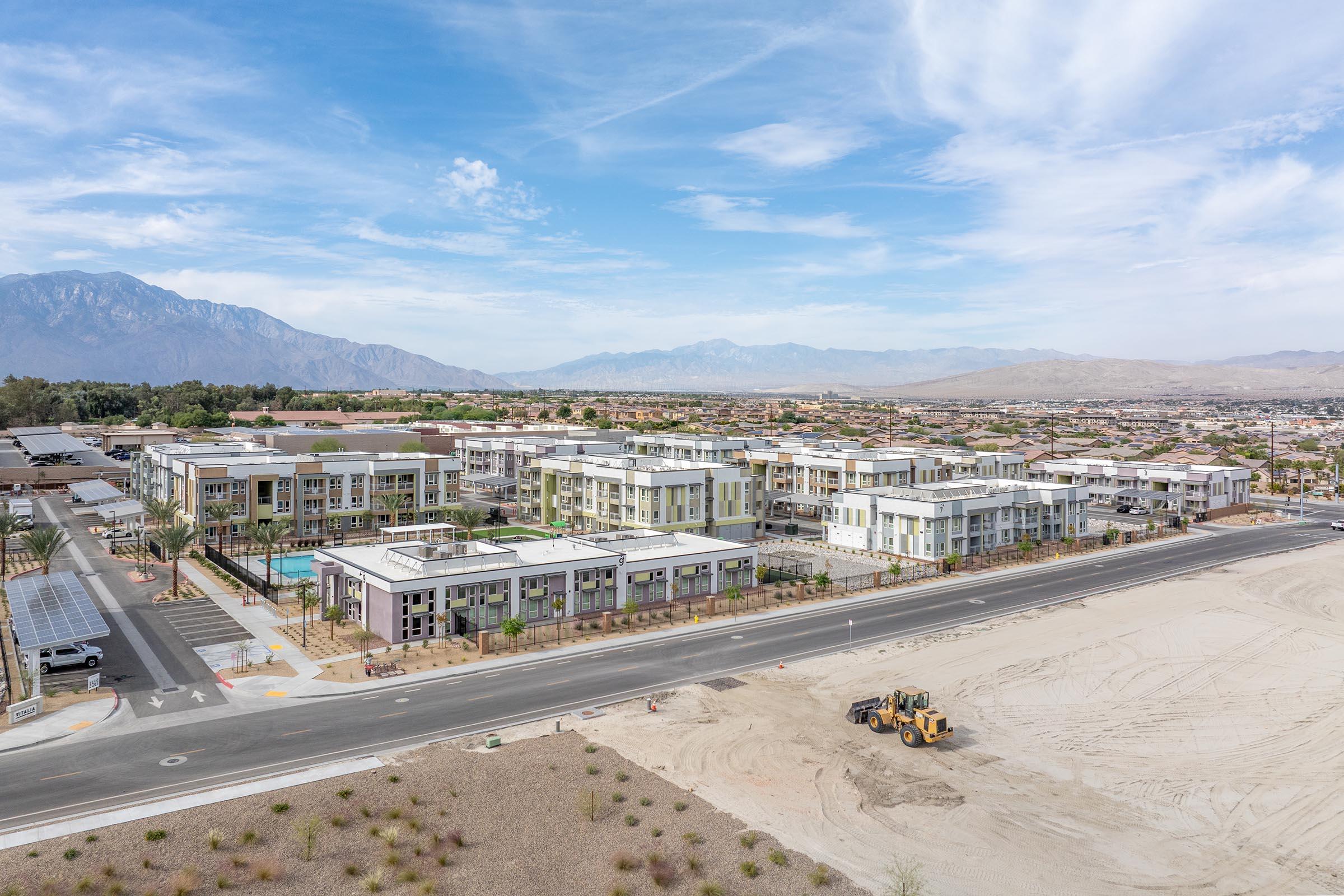 Aerial view of a modern apartment complex with several buildings, a swimming pool area, and surrounding desert landscape. In the foreground, a construction vehicle is parked on a sandy area. The background features distant mountains under a partly cloudy blue sky.