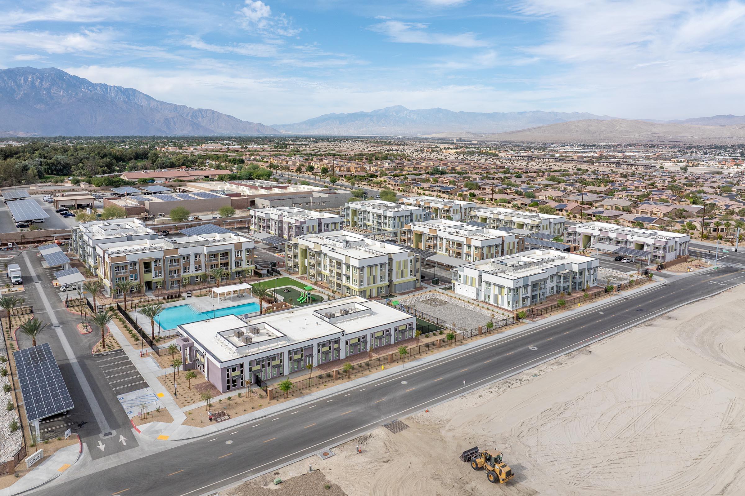 Aerial view of a modern apartment complex featuring multiple buildings with flat roofs, a pool, and landscaped areas. Surrounding the complex are residential neighborhoods and mountains in the background. An empty lot and a road are visible in the foreground, indicating ongoing development in the area.