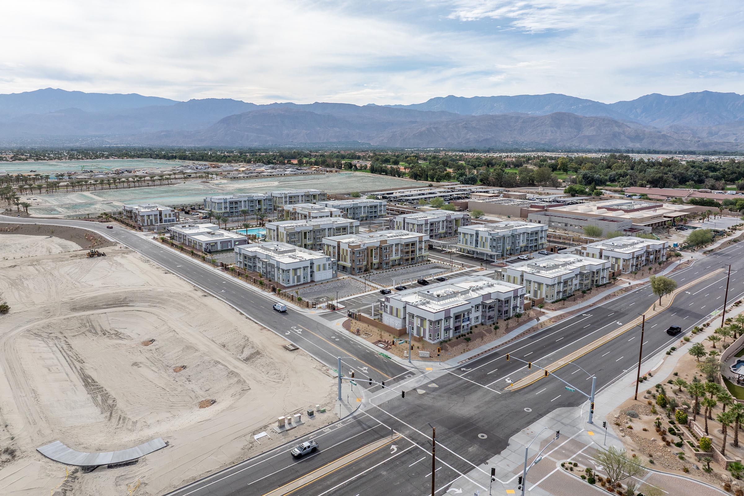 Aerial view of a new residential development with several modern buildings under construction. The area features roads, landscaping, and mountain scenery in the background. Dusty land indicates ongoing development, while some vehicles can be seen on the roads. Overall, the scene captures a suburban expansion.