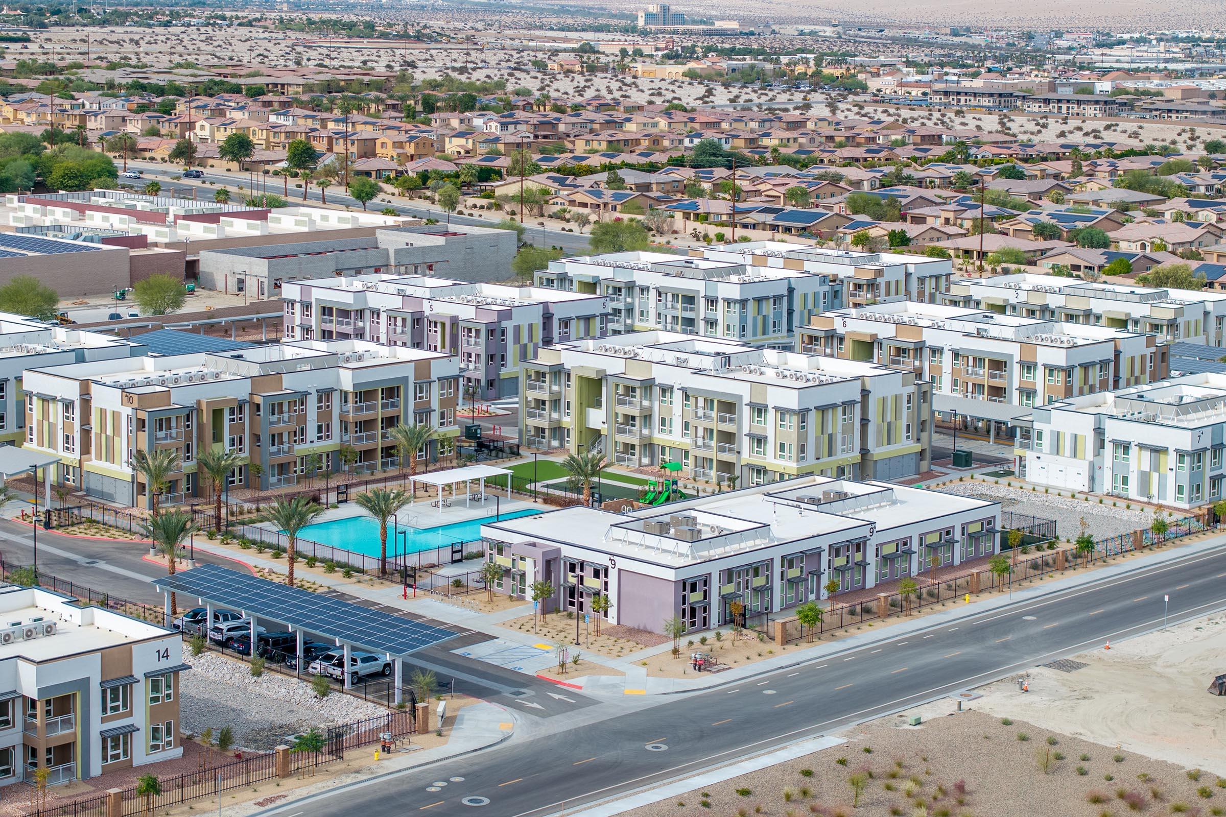 Aerial view of a modern residential complex featuring several multi-story buildings with swimming pool and palm trees. Surrounding area includes other houses and neighborhoods, with a backdrop of desert landscape and distant mountains. Clear skies above and well-maintained landscaping around the buildings.