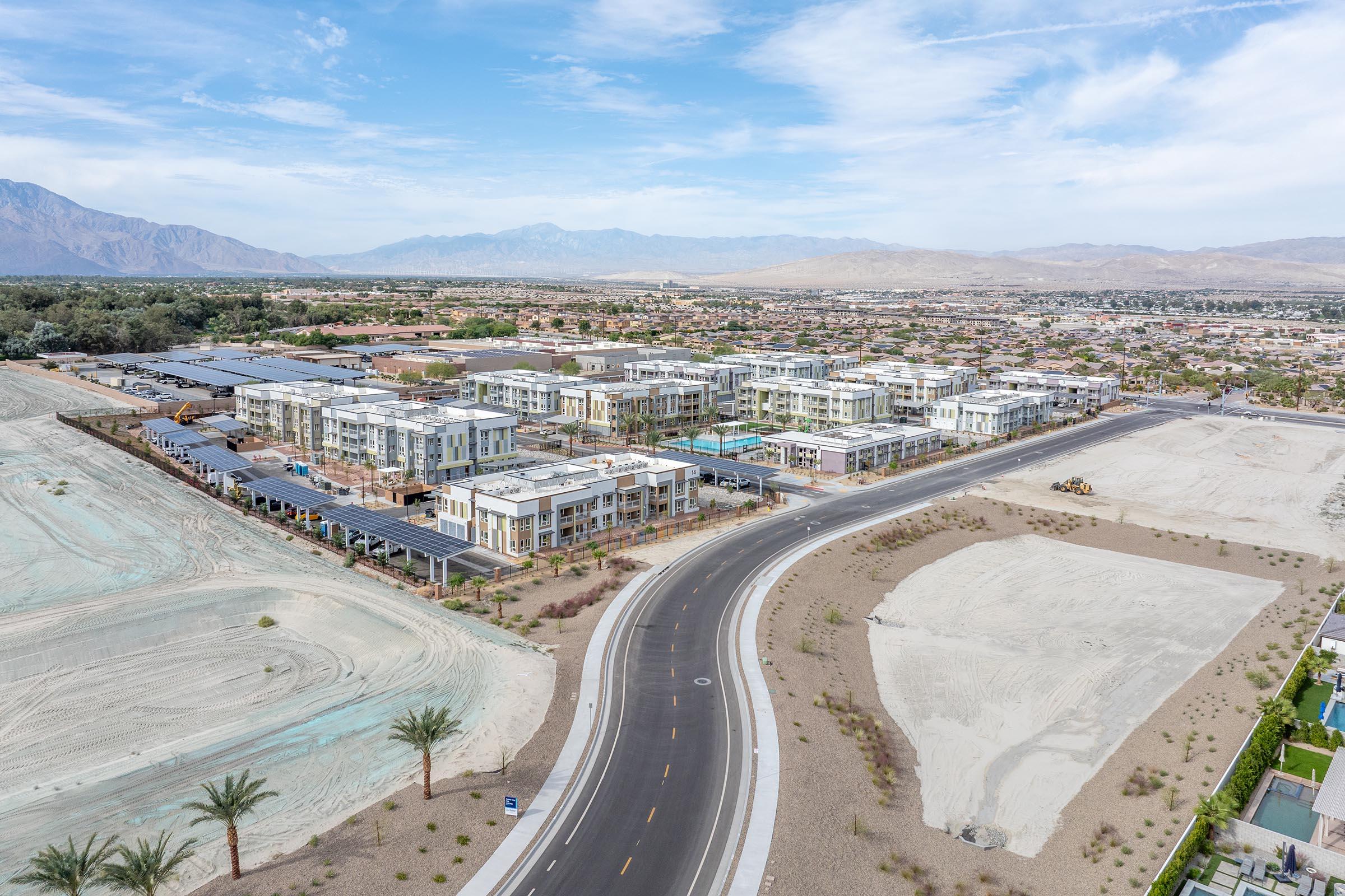 Aerial view of a partially developed residential area with modern buildings and a curved road. Surrounding the development are empty lots and desert landscape with mountains in the background. The sky is partly cloudy, and there are palm trees lining the road.