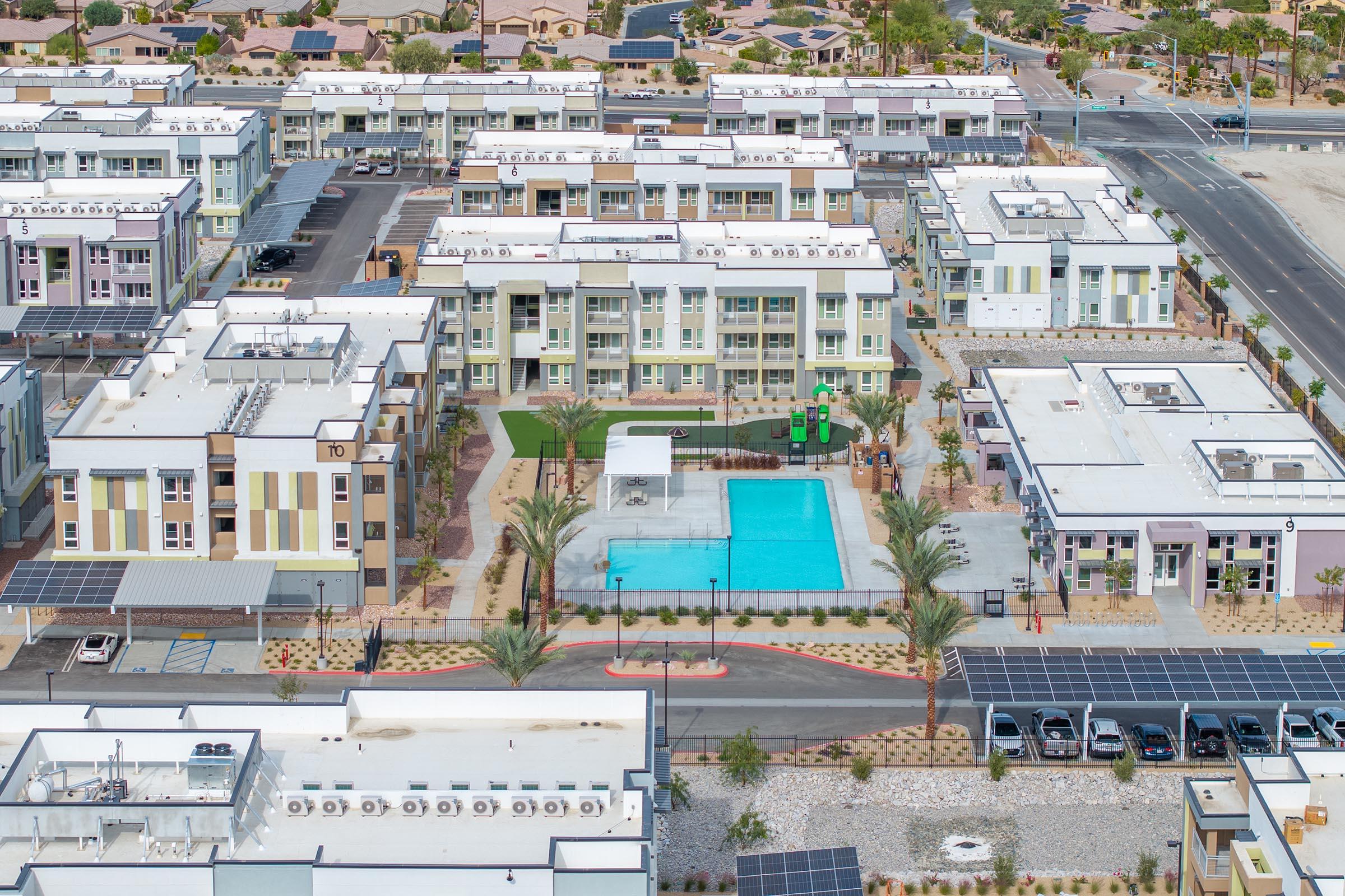 Aerial view of a modern apartment complex featuring multiple buildings with a central swimming pool and landscaped areas. Solar panels are visible on rooftops, and surrounding infrastructure includes parking lots and pathways. The area appears well-maintained and is set in a suburban environment.