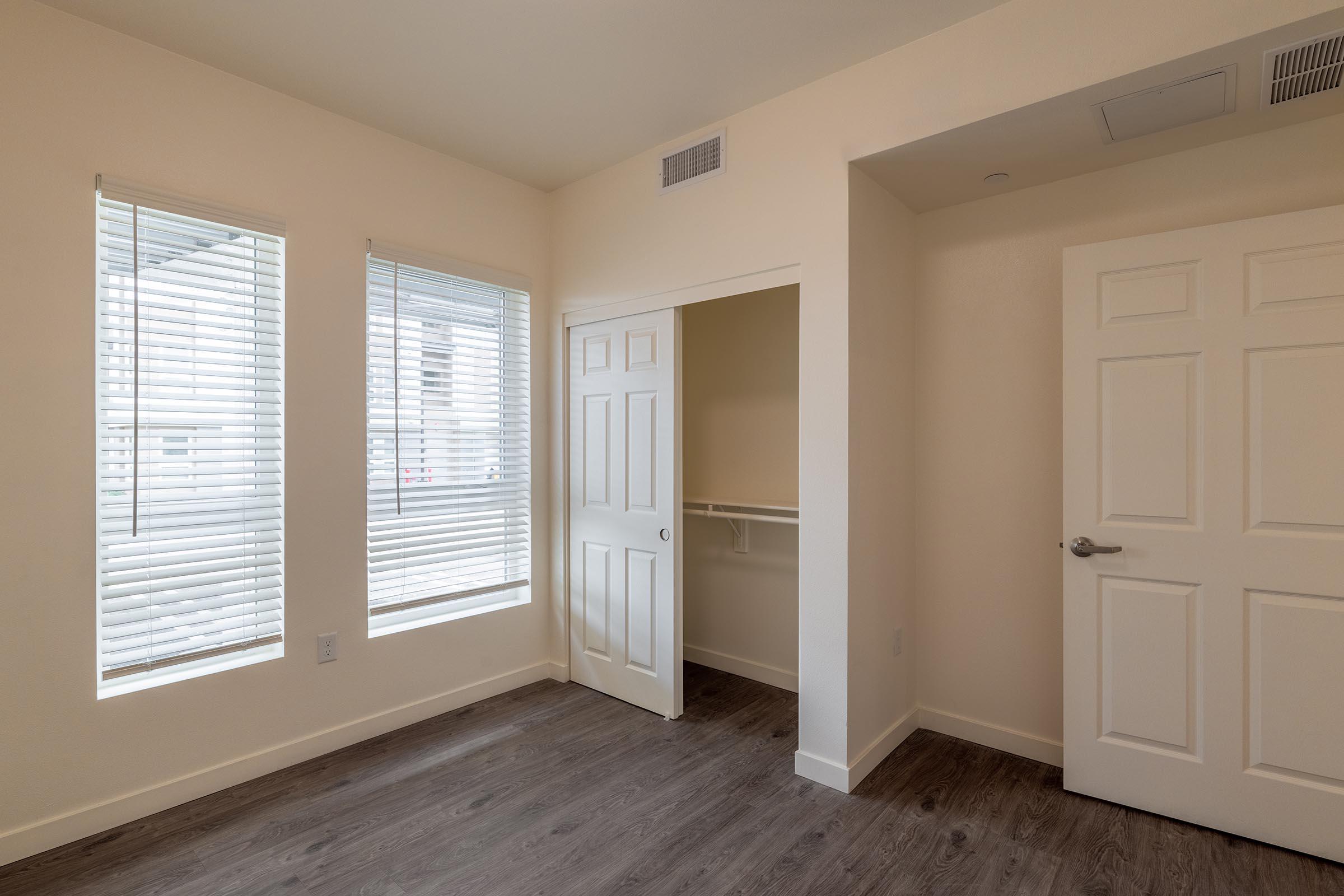 A well-lit, empty room featuring two windows with white blinds, light-colored walls, and a hardwood floor. There is a closet with an open door on the right side, and a solid door leading to another space. The overall ambiance is clean and modern, suitable for various uses.