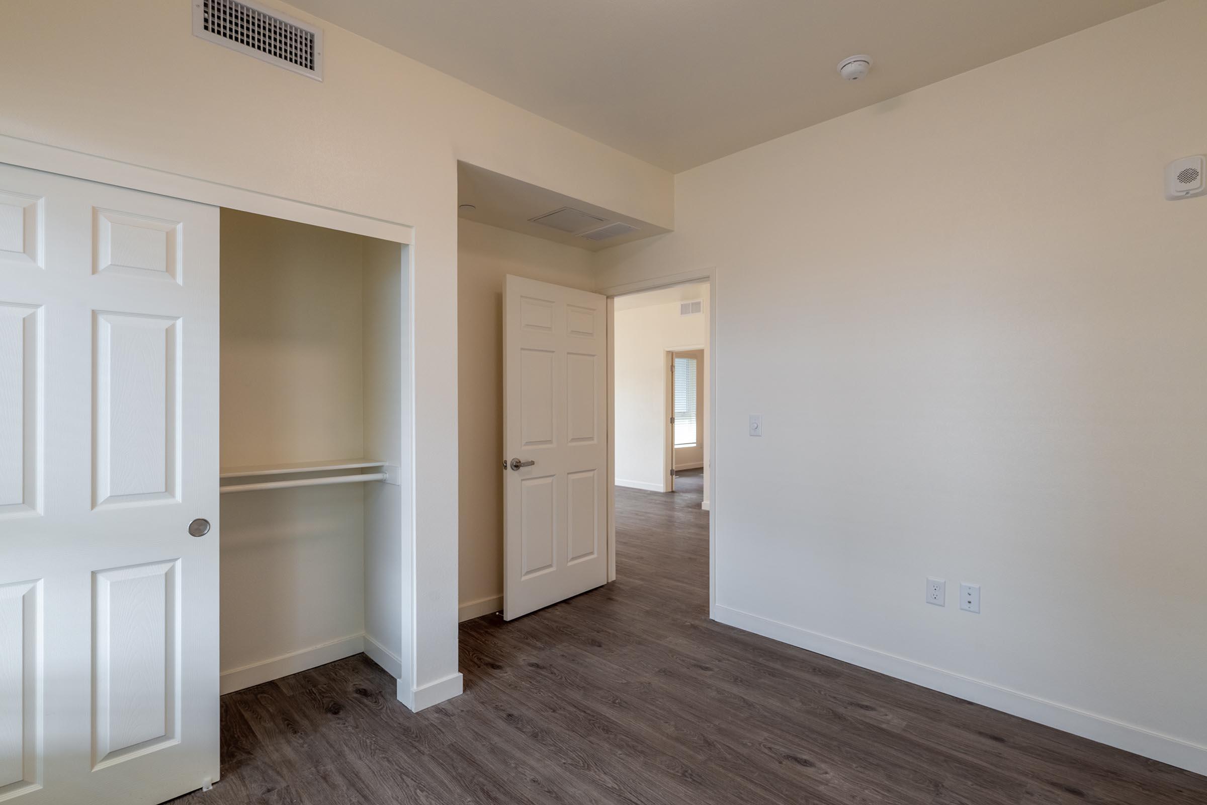 Interior view of a room featuring light-colored walls and wood-look flooring. There is a doorway leading to another room, and a closet with double doors on the left. The space is well-lit with a neutral aesthetic, creating an open and inviting atmosphere.