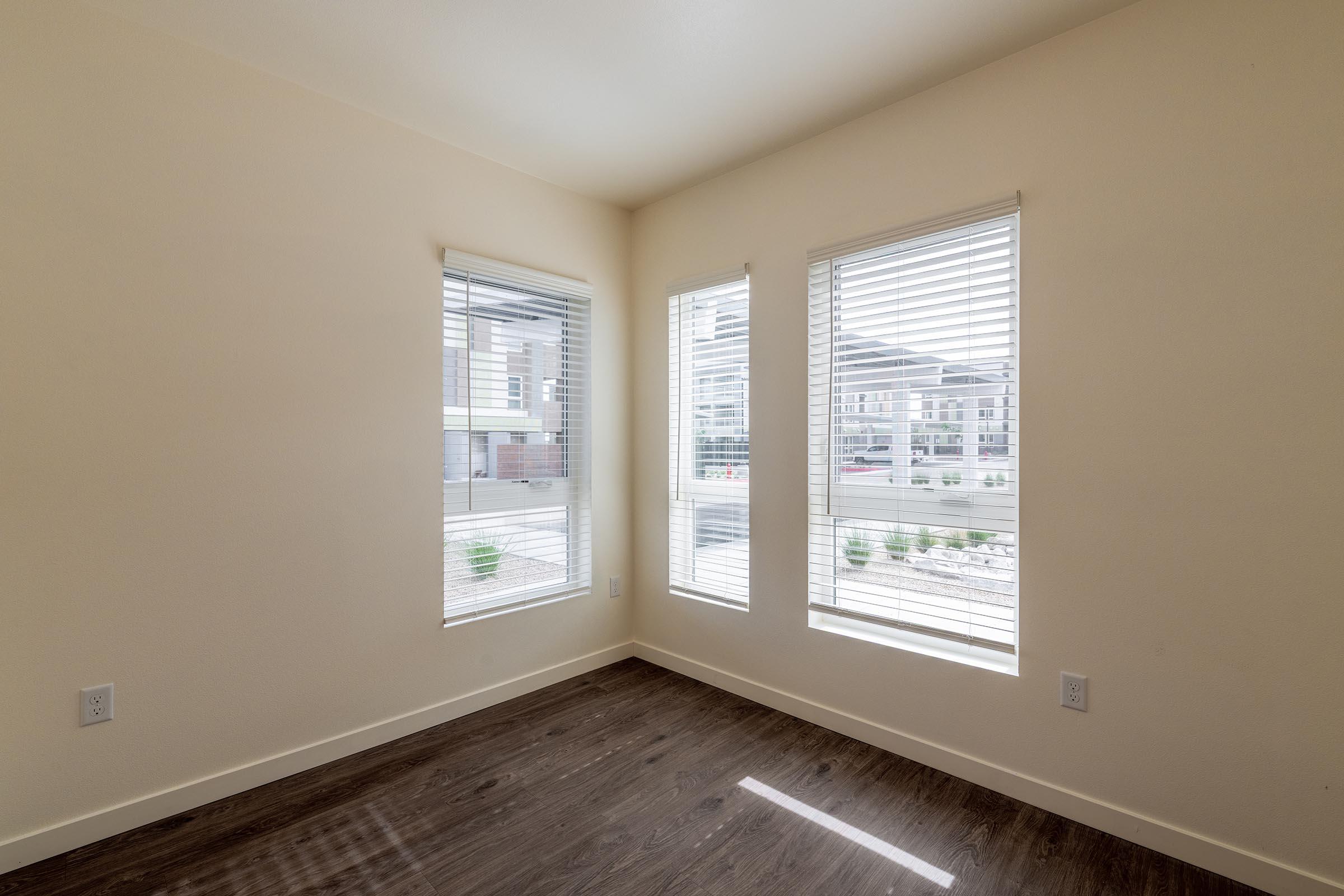 A bright, empty room with light-colored walls and dark wood laminate flooring. Three windows with blinds allow natural light to enter, revealing a view of the outside. The space is clean and unadorned, suitable for various uses.